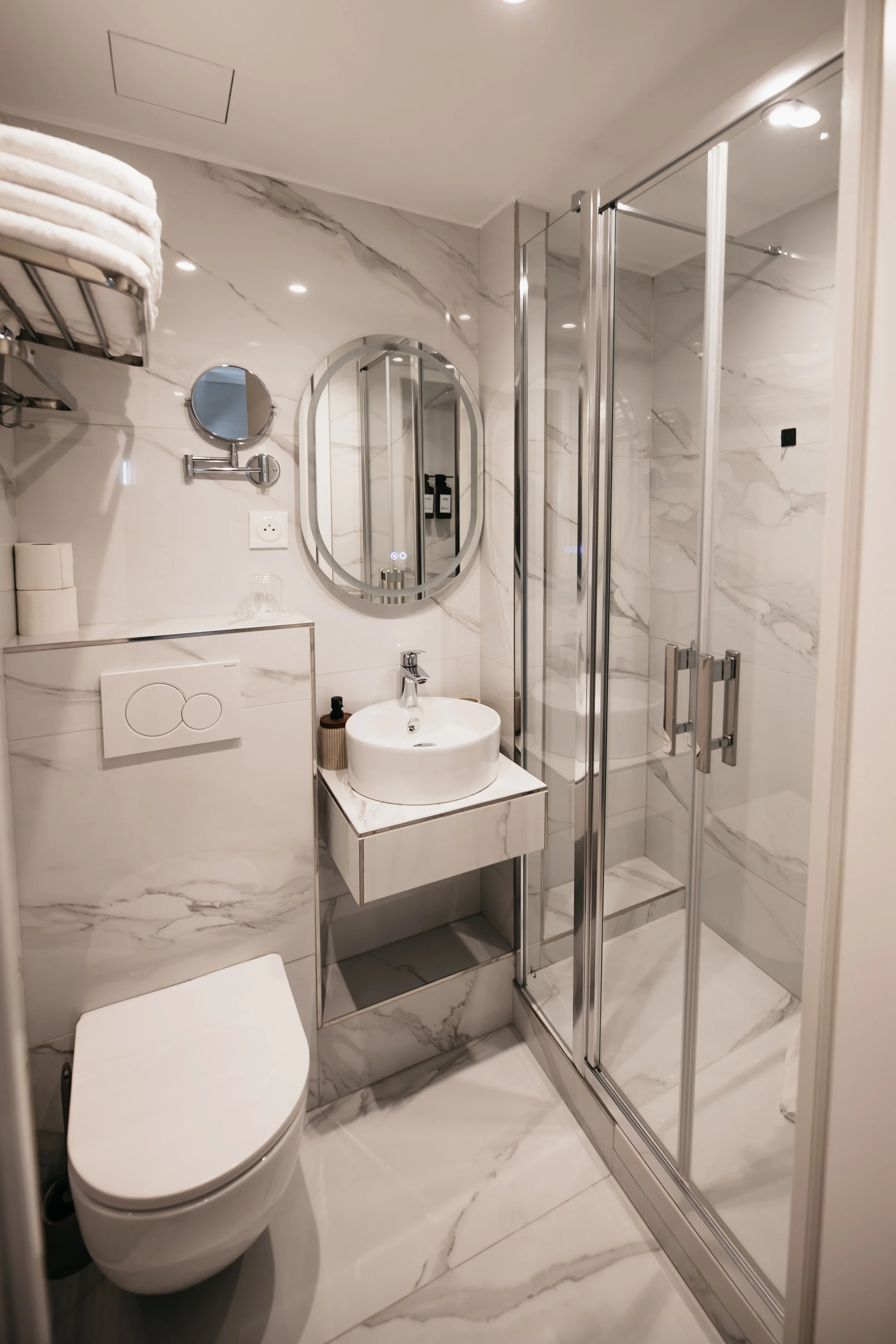 Modern bathroom with white marble walls and floor, a white toilet, small white sink with a mirror above, glass shower enclosure with chrome fixtures, towel rack with folded towels, and toiletries arrangements.