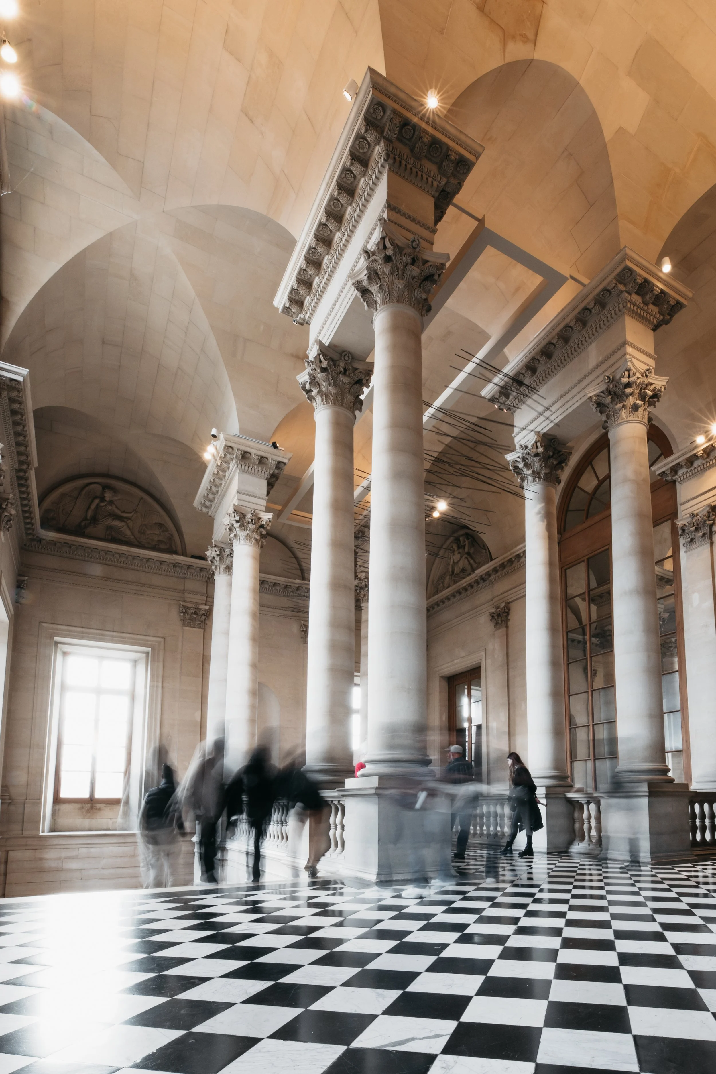 Interior of a grand hall with tall classical columns with ornate capitals, checkered black-and-white floor, high arched ceilings, and large windows allowing natural light. People are walking through, creating a blurred motion effect.