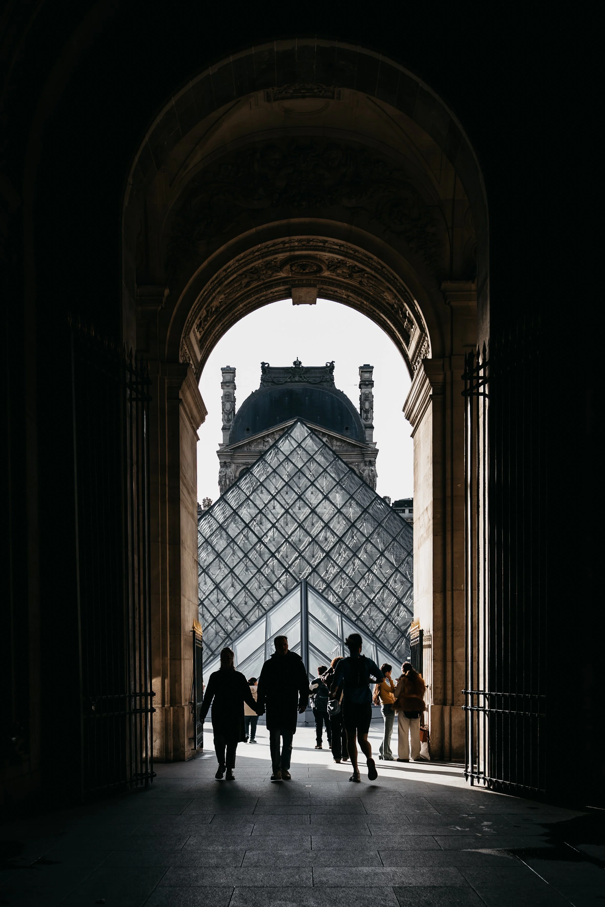 People walking through an arched gateway towards the glass and metal Louvre Pyramid with historical building in the background. photography made in December 