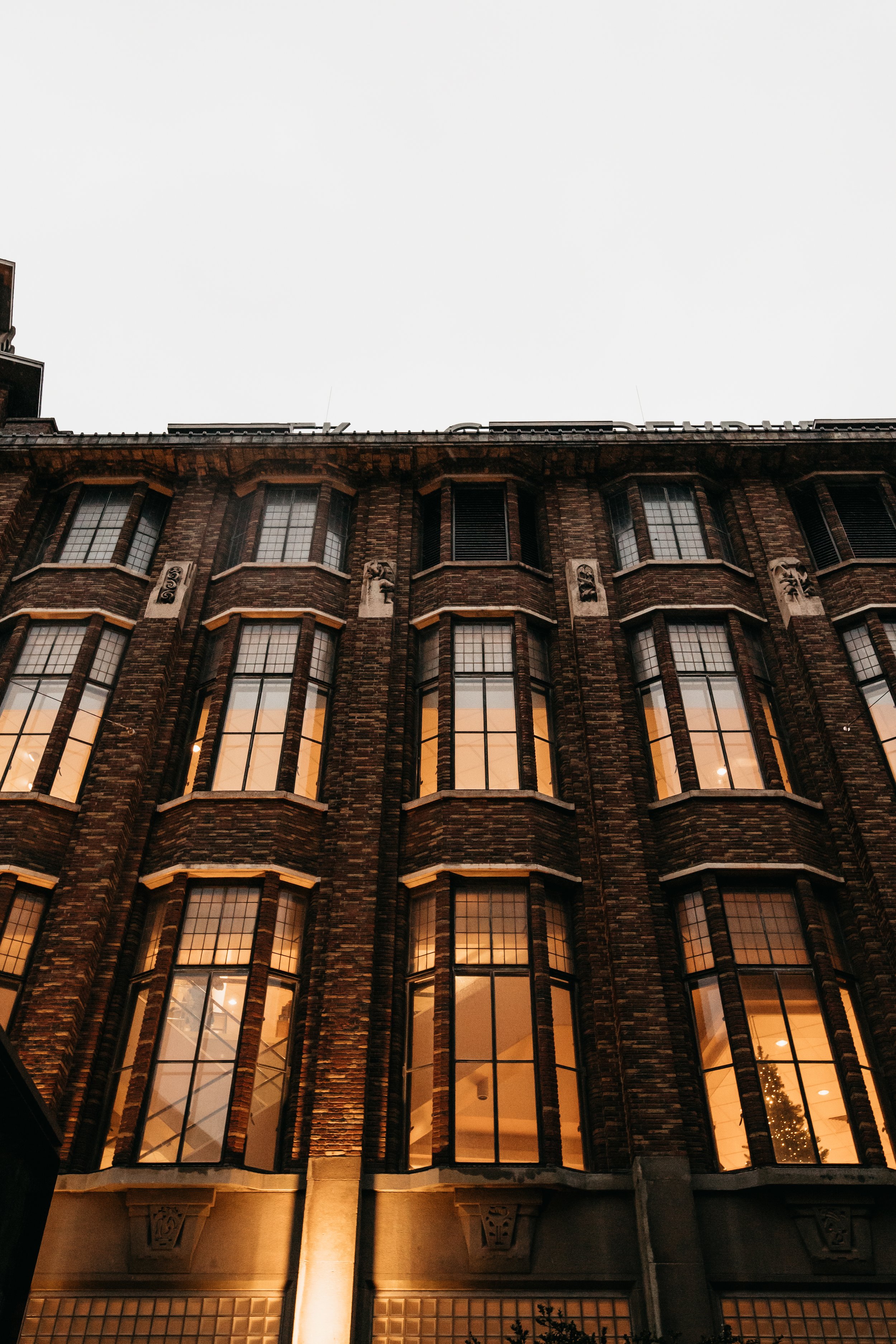 A multi-story brick building with large windows warm interior lighting, and some decorative architectural details on the facade. Made in The Hague with canon 6D. 