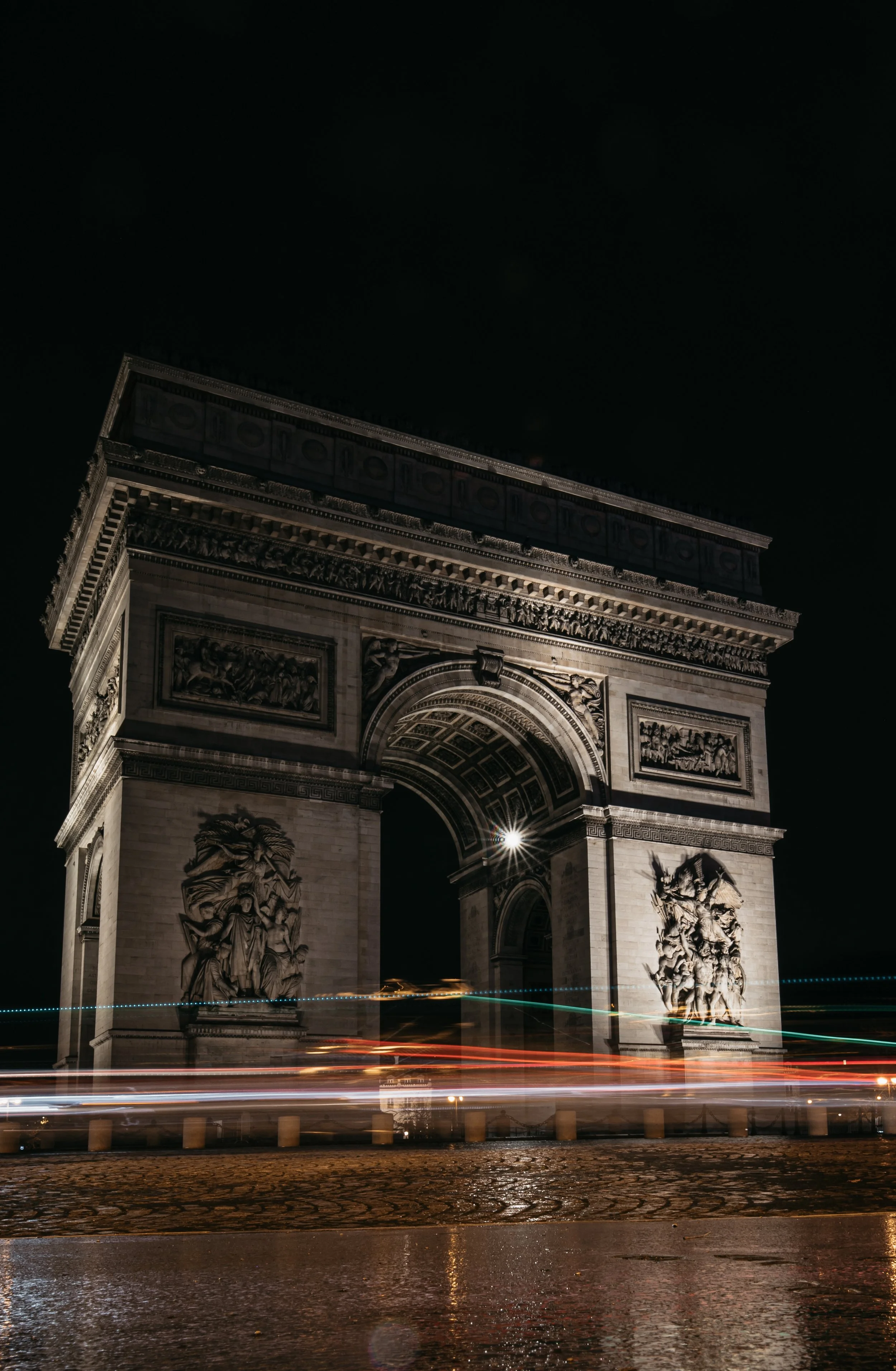 Nighttime view of the Arc de Triomphe in Paris illuminated, with light trails from moving vehicles on wet pavement in the foreground.