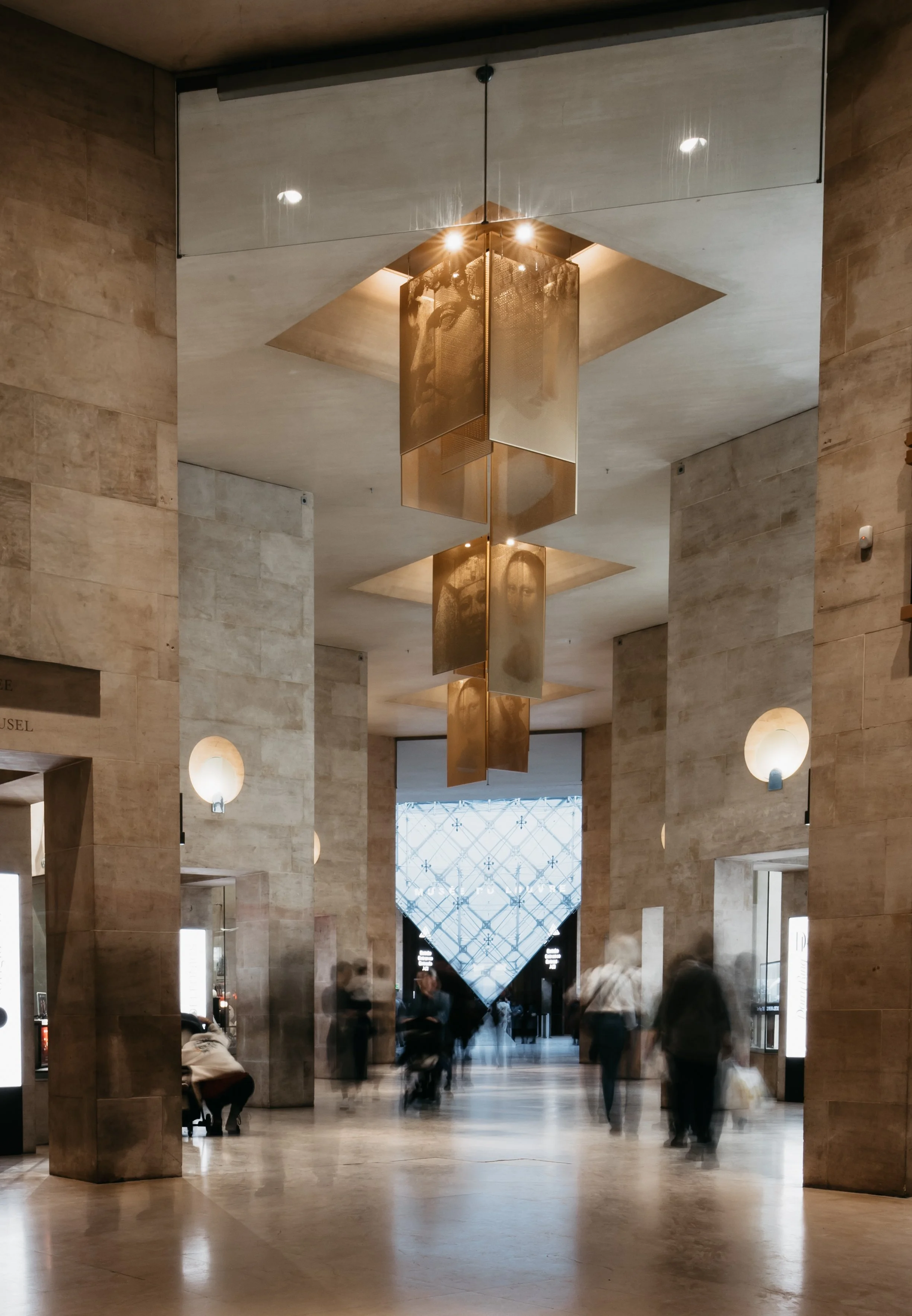 Interior of a modern building with a high ceiling, blurred people walking, a large glass entrance, and a decorative hanging light fixture.
