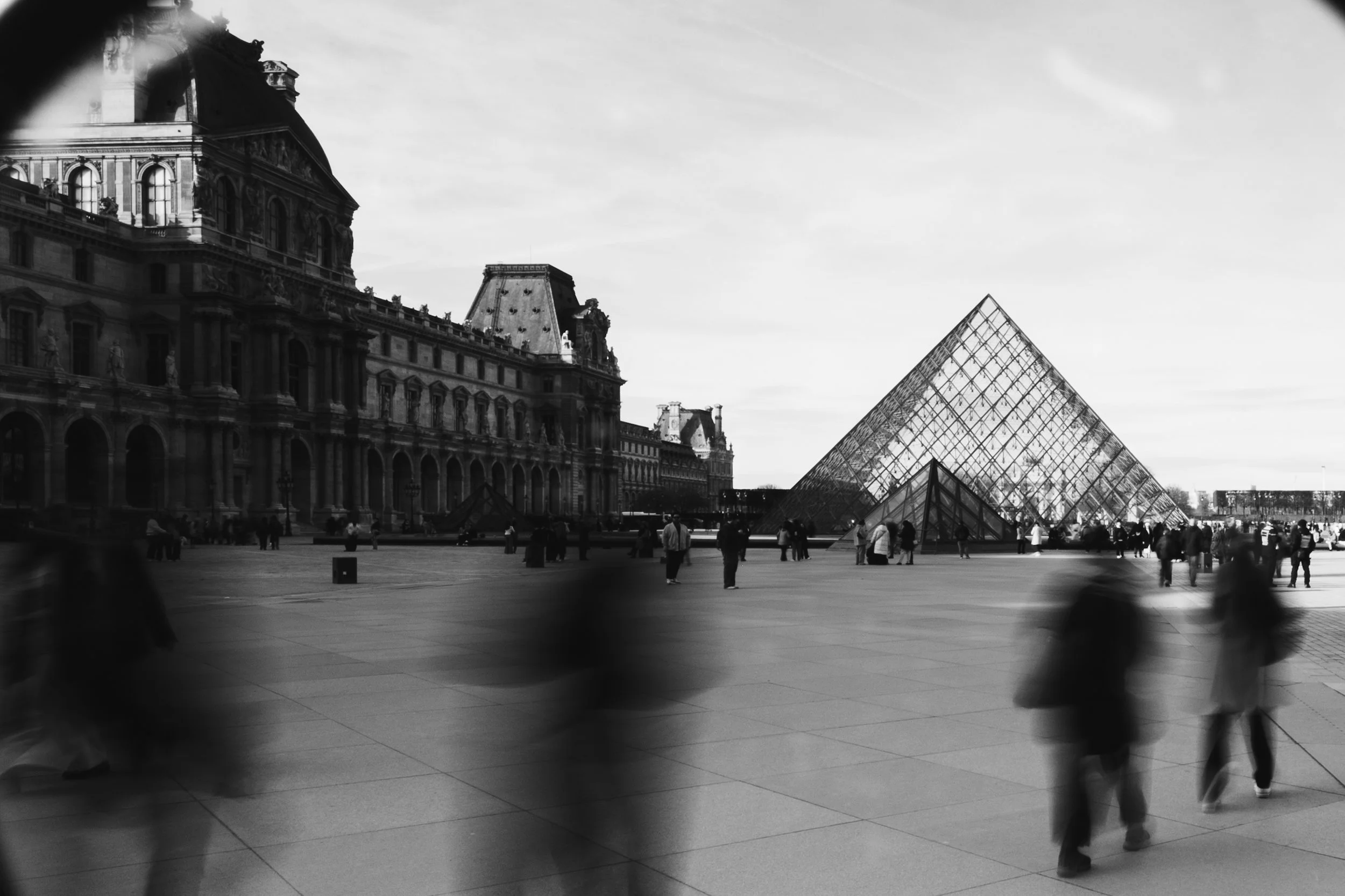 Black and white photo of the Louvre Museum and glass pyramid in Paris, with people walking and standing in the courtyard.