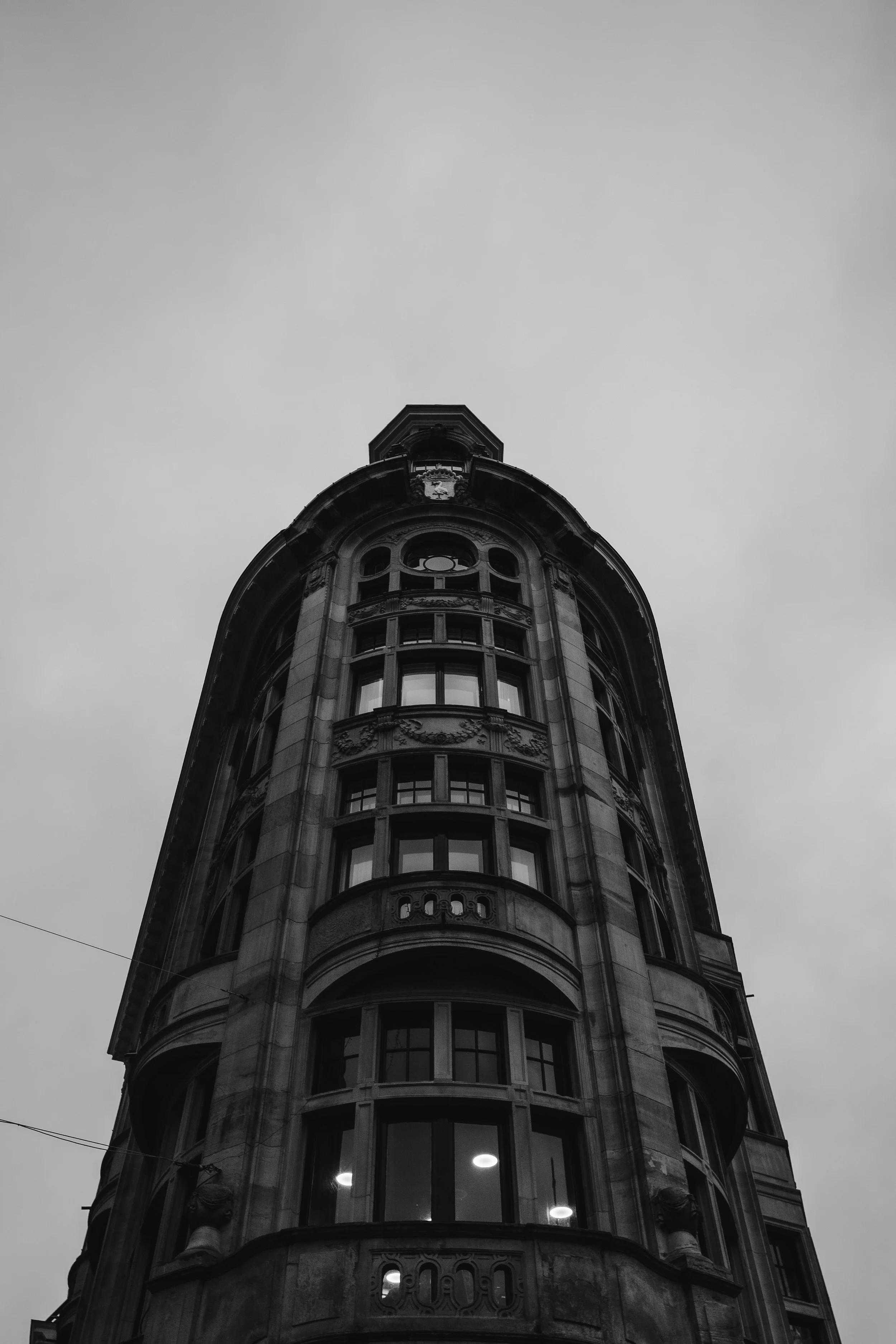 A black and white photo of a tall, historic building with ornate architectural details and multiple windows, viewed from below with a cloudy sky in the background.