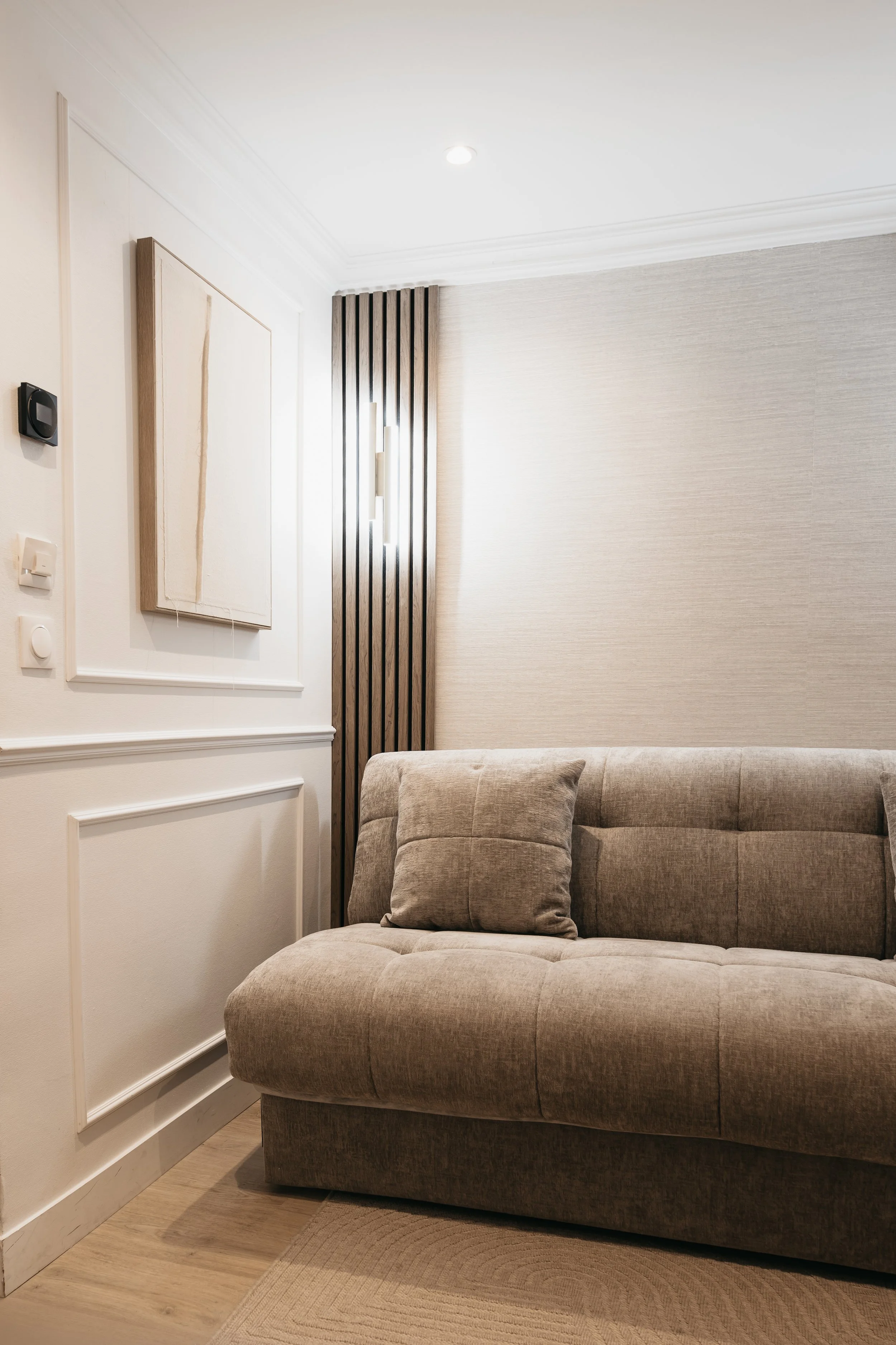 Modern living room corner with beige sofa, cushion, abstract wall art, decorative wood panel, wall sconce, and textured wallpaper.