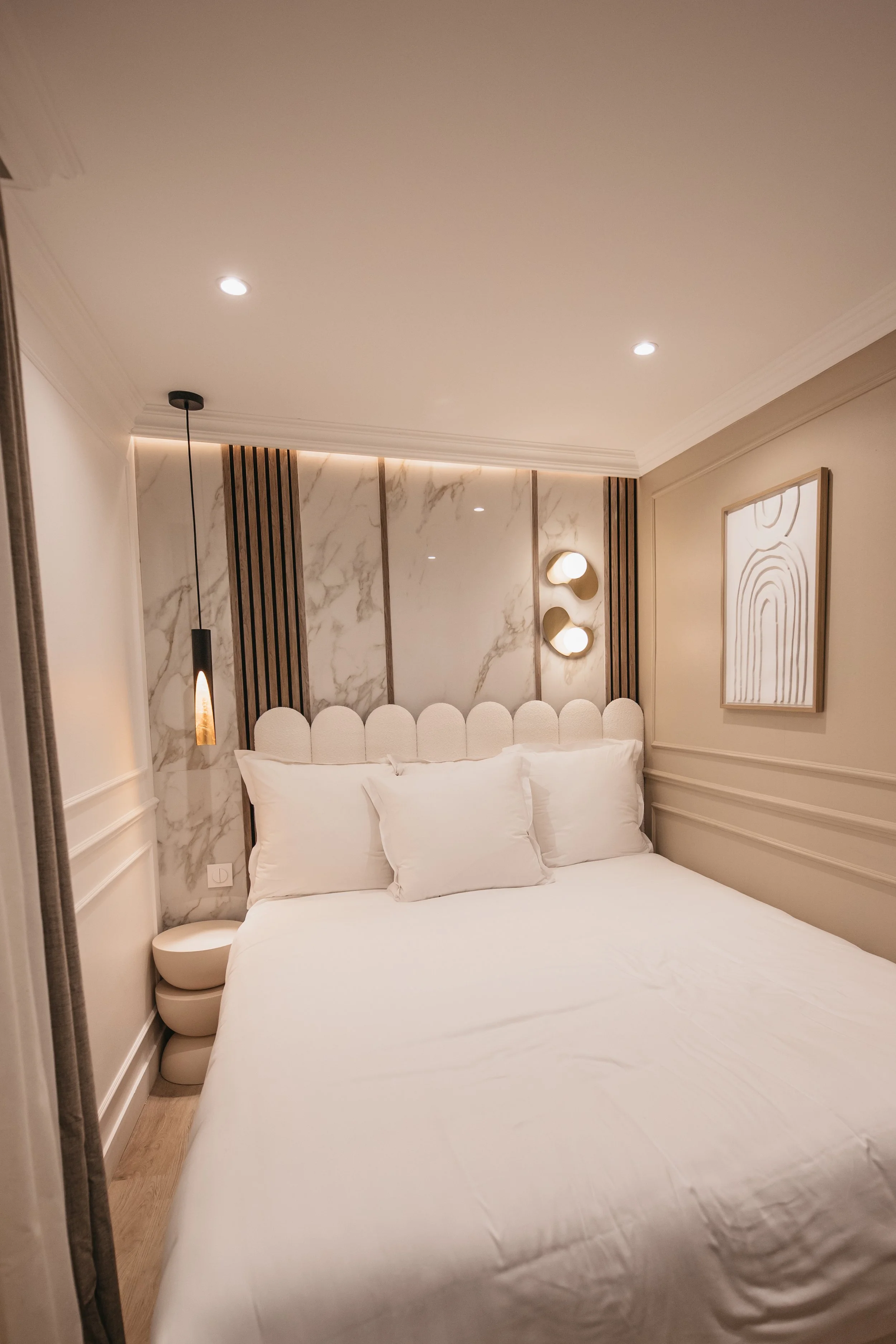 A modern bedroom with a bed, white bedding, and pillows, a marble accent wall behind the bed, and contemporary hanging and wall-mounted light fixtures. interior photography in a hotel in Paris