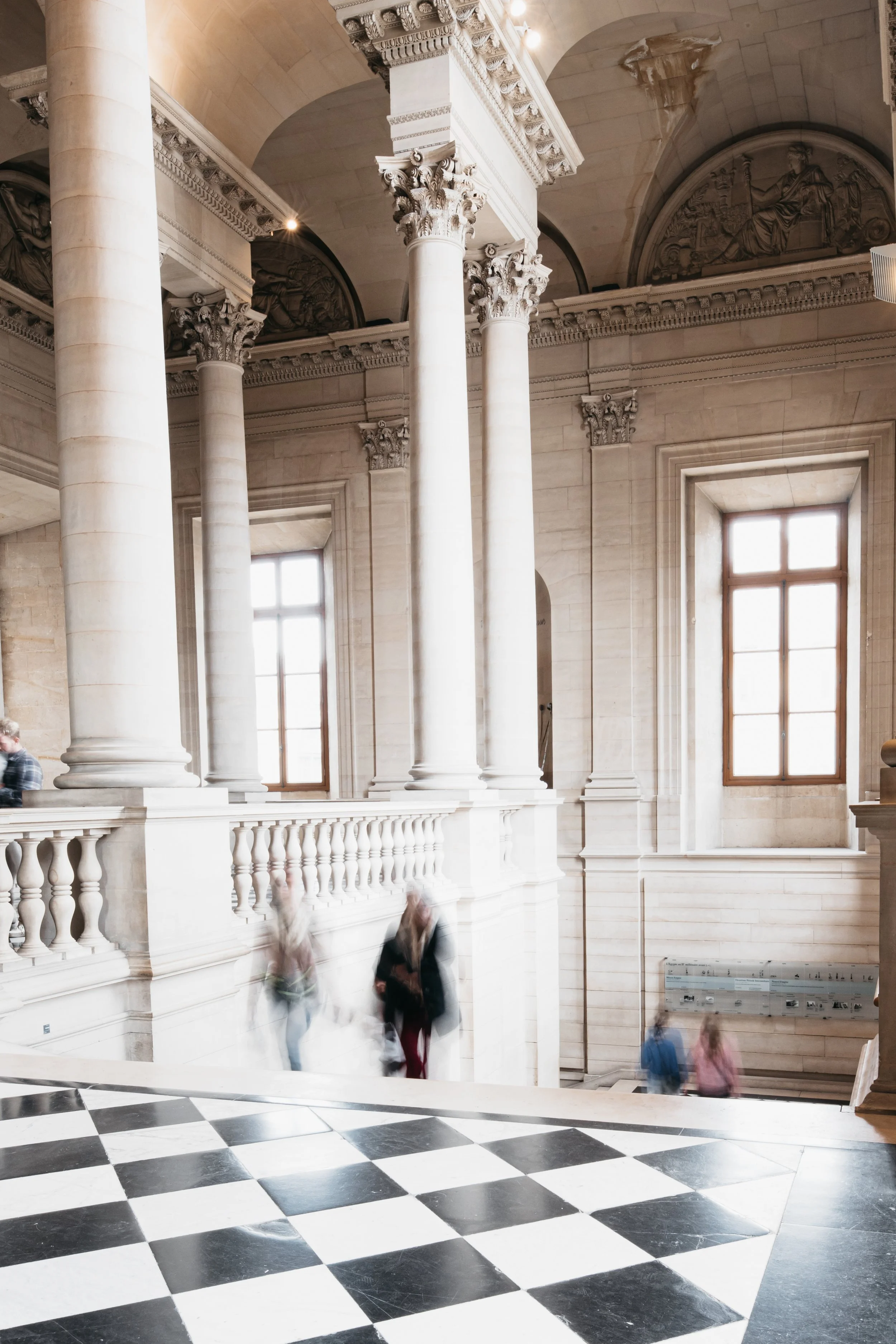 Interior of a historic building with tall white columns, large windows, and a black-and-white checkered floor. Some blurred people are walking through the space. Louvre - Paris photography