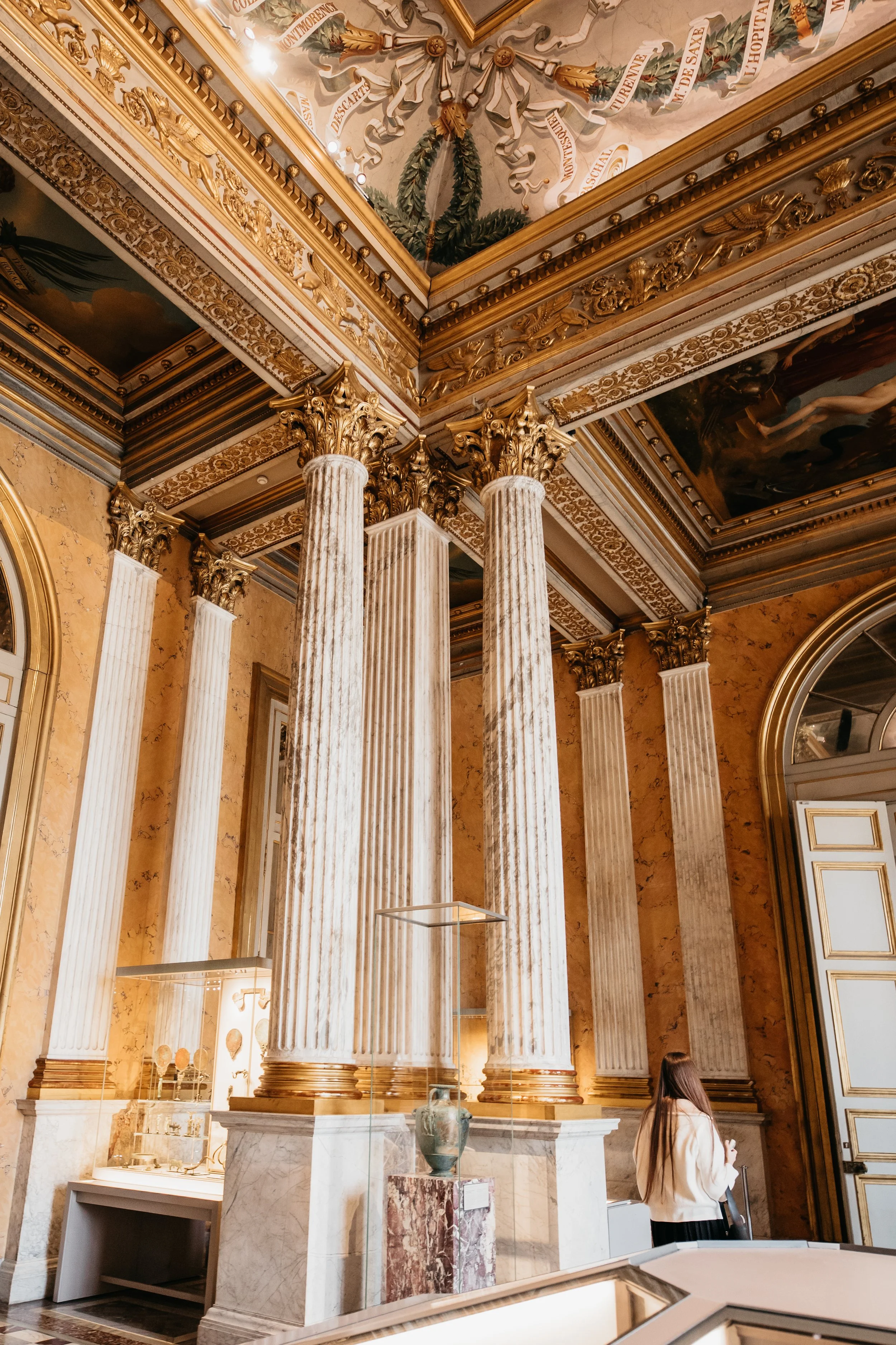 Interior of an ornate museum with four tall marble columns with gold accents, detailed ceiling artwork, and a woman observing the exhibits.