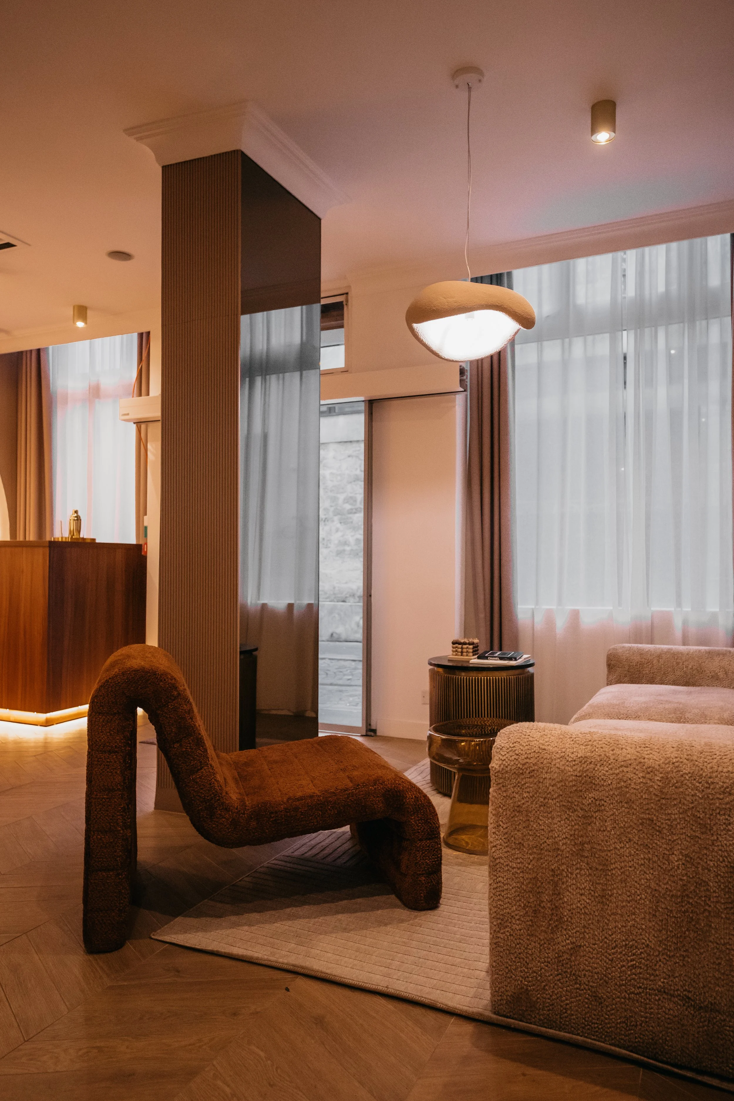 Interior of a modern living room featuring a brown chair, a beige sofa, a round side table, a pendant light, and large windows with white curtains. interior photography made in a hotel in Paris
