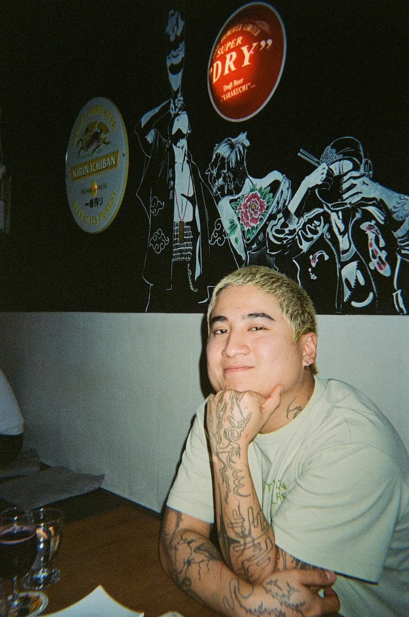 Jeune homme avec des tatouages assis à une table dans un restaurant, avec un mur décoratif en arrière-plan représentant des personnages de style manga ou graffiti, certains tenant des boissons ou des accessoires.