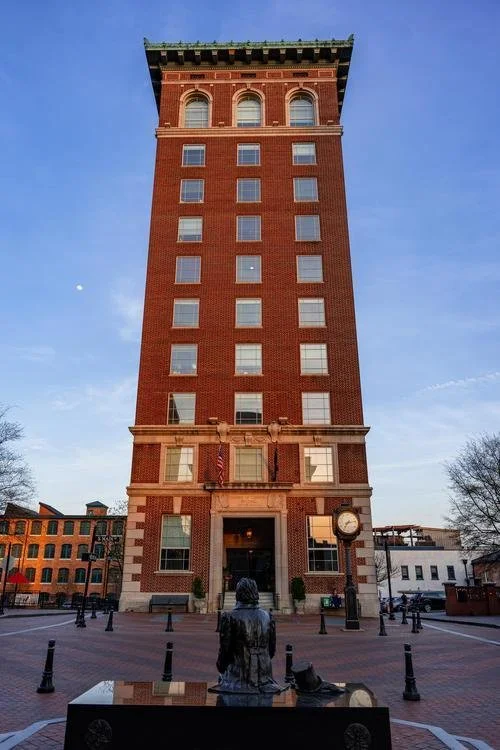 A tall red brick clock tower with windows and a clock near the entrance, situated in a small town square with a person sitting on a bench in the foreground and other brick buildings nearby, under a clear blue sky.