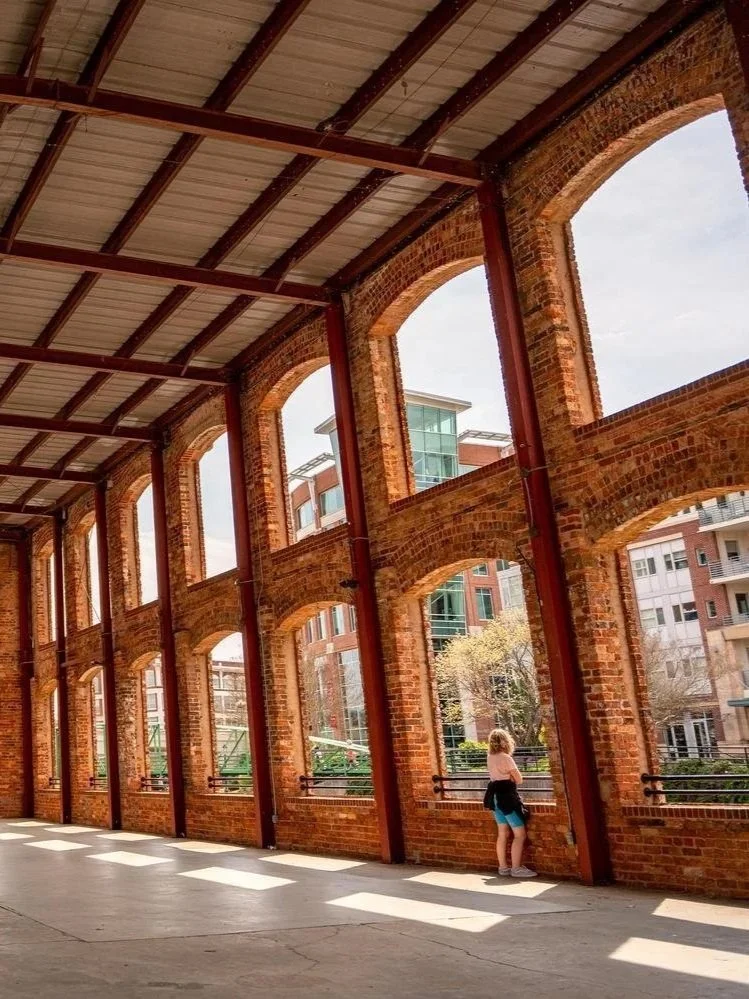 A person stands inside a brick building with large arched windows, looking out at a cityscape with modern buildings and trees.