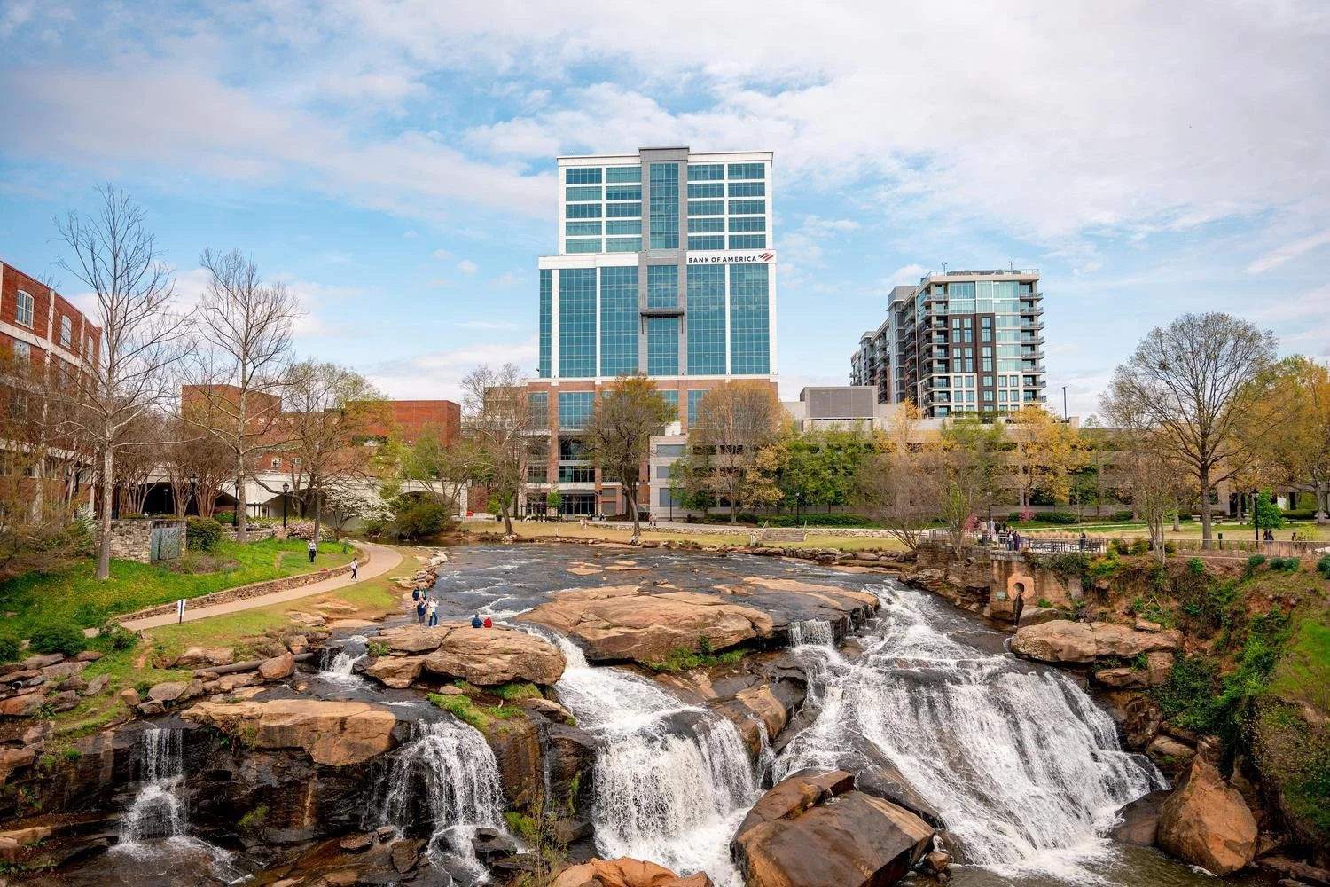 City park with a waterfall and walking path in front of modern high-rise buildings, trees, and a blue sky with clouds.