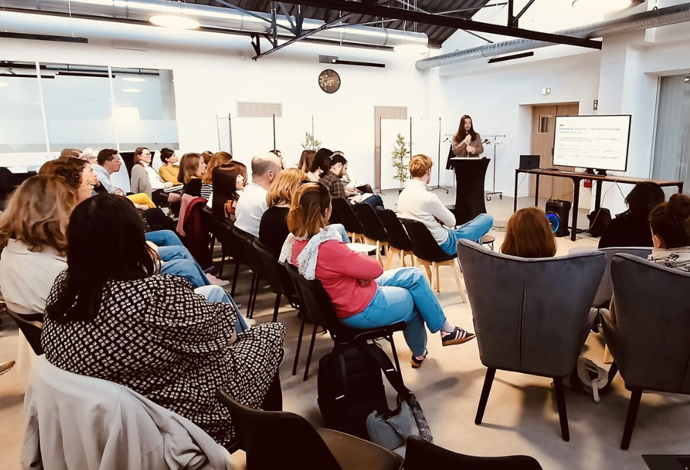 Une salle de conférence avec une femme donnant une présentation devant un écran et un public de plusieurs personnes assises, écoutant attentivement.