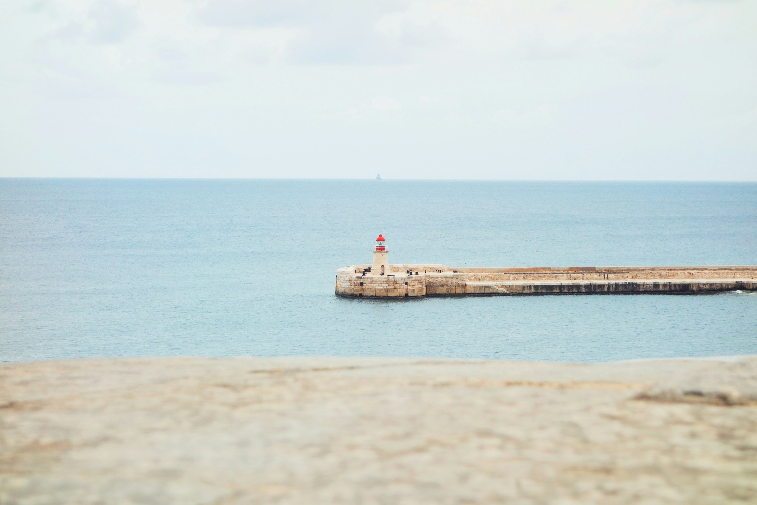 Un phare rouge et blanc situé sur une jetée en pierre dans la mer, avec un ciel nuageux et une silhouette de bateau à l'horizon.