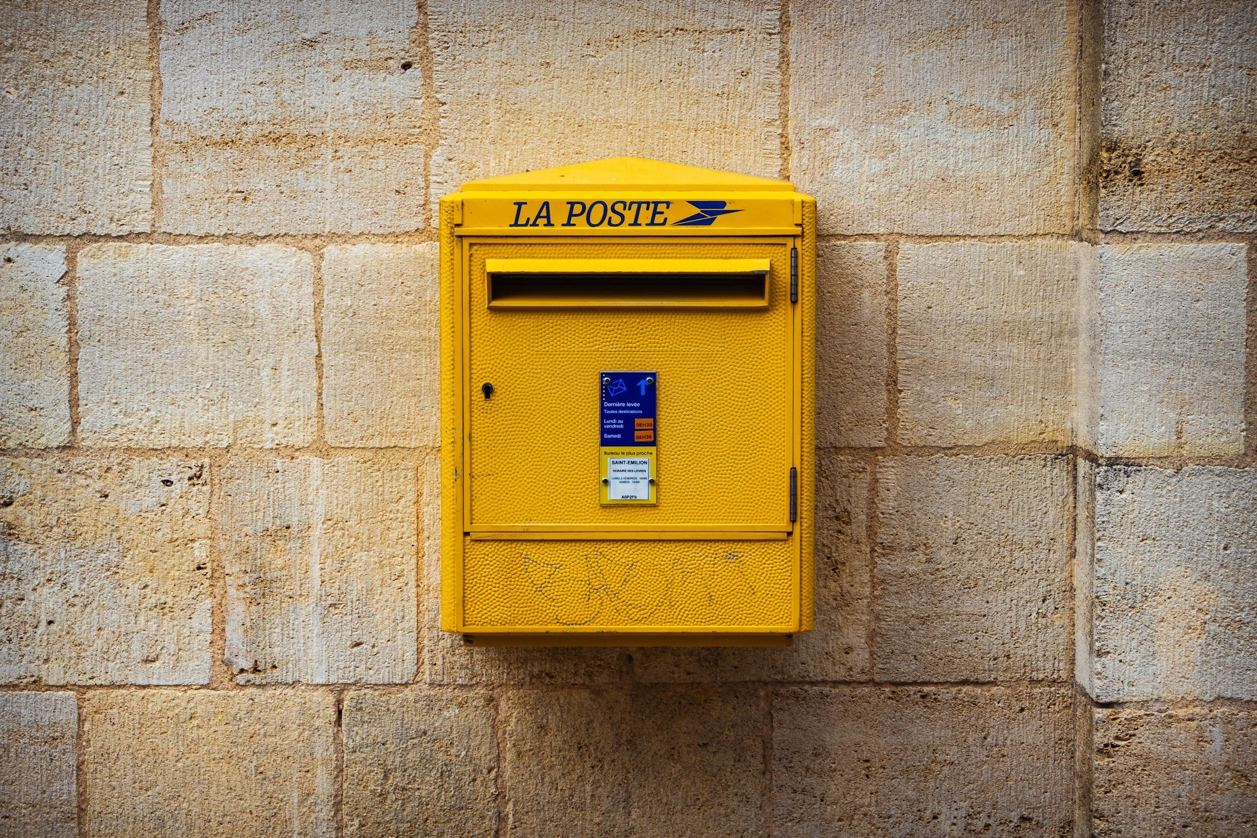 Boîte aux lettres jaune fixée à un mur en pierre, avec l'inscription 'LA POSTE' en bleu.