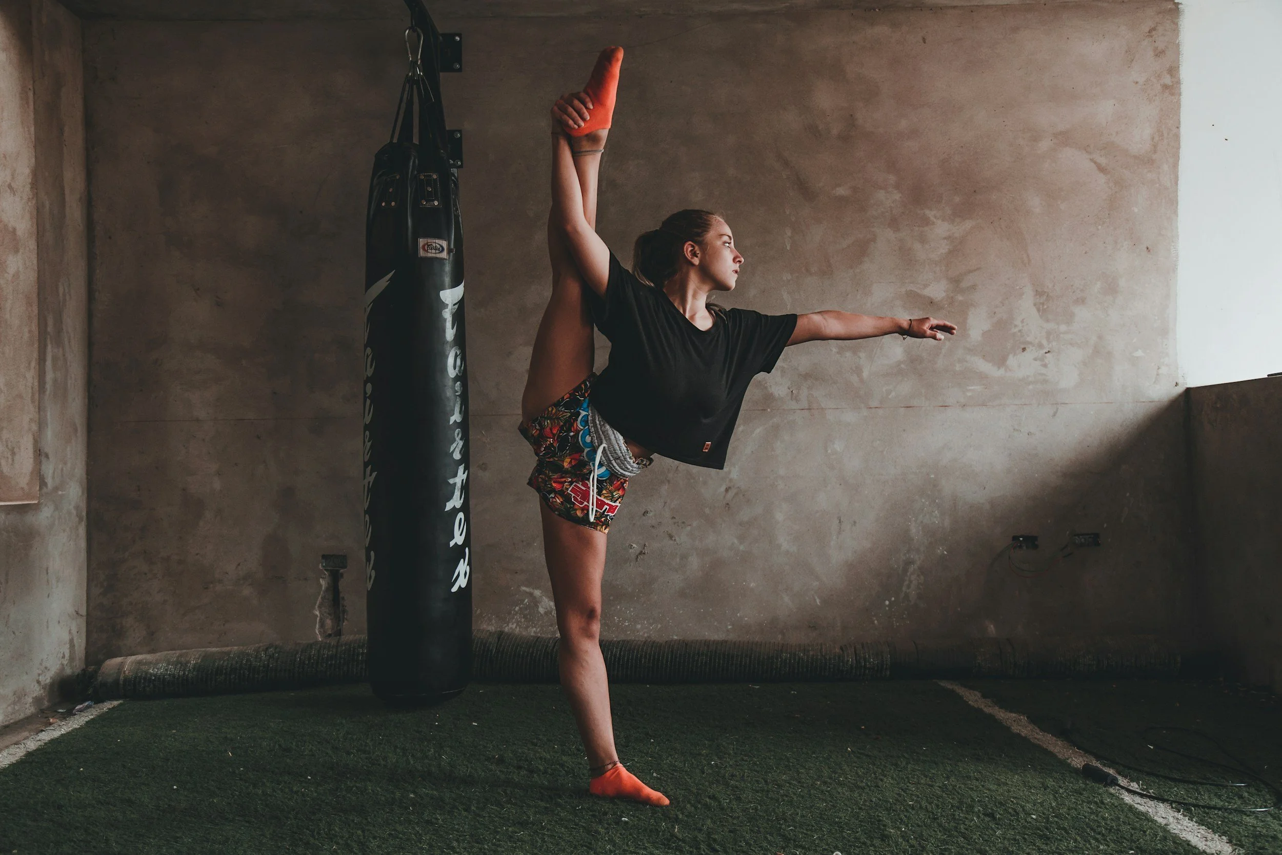 Jeune femme faisant de la poses d'étirement dans une salle de sport avec un sac de boxe suspendu et une surface de sol en gazon artificiel.