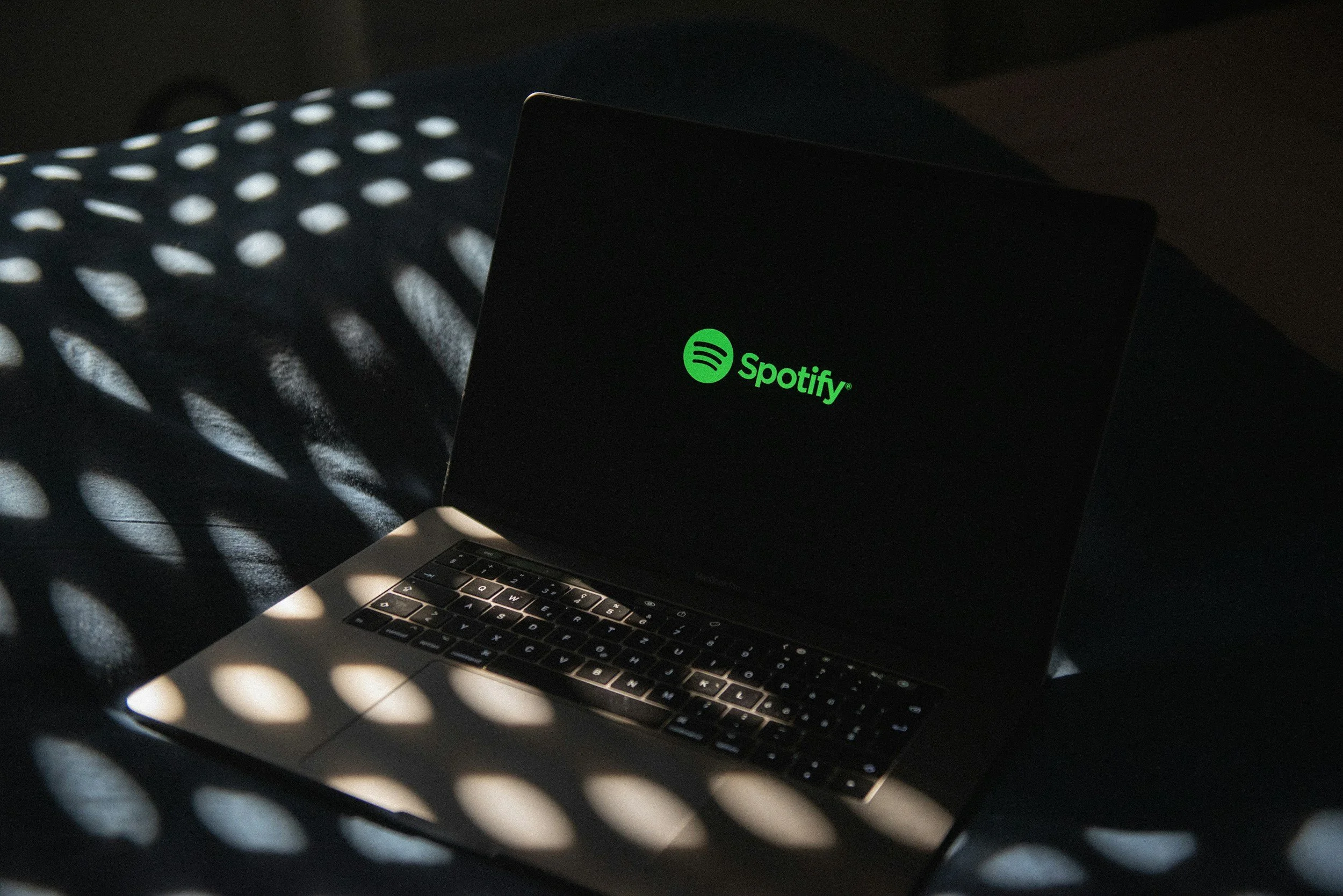 Ordinateur portable avec le logo Spotify sur l'écran, placé sur une surface sombre avec des motifs de lumière et d'ombre créés par des objets perforés.