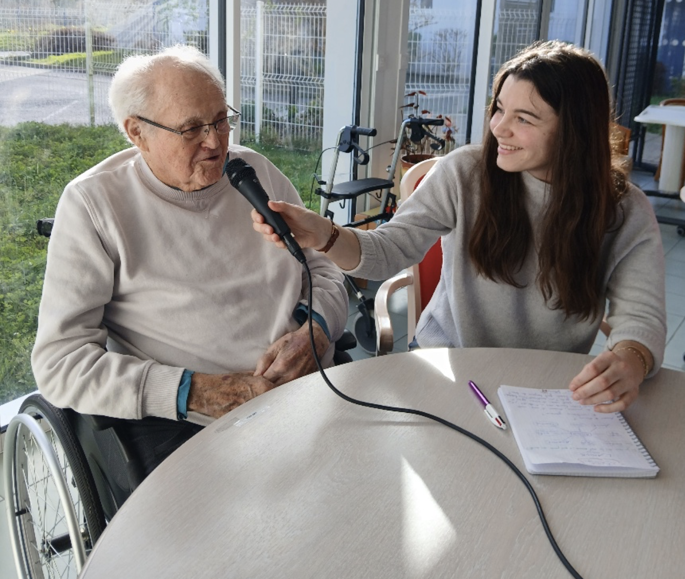 Une jeune femme interviewant un homme âgé en fauteuil roulant dans une pièce lumineuse avec de grandes fenêtres, un carnet et un stylo sur la table.