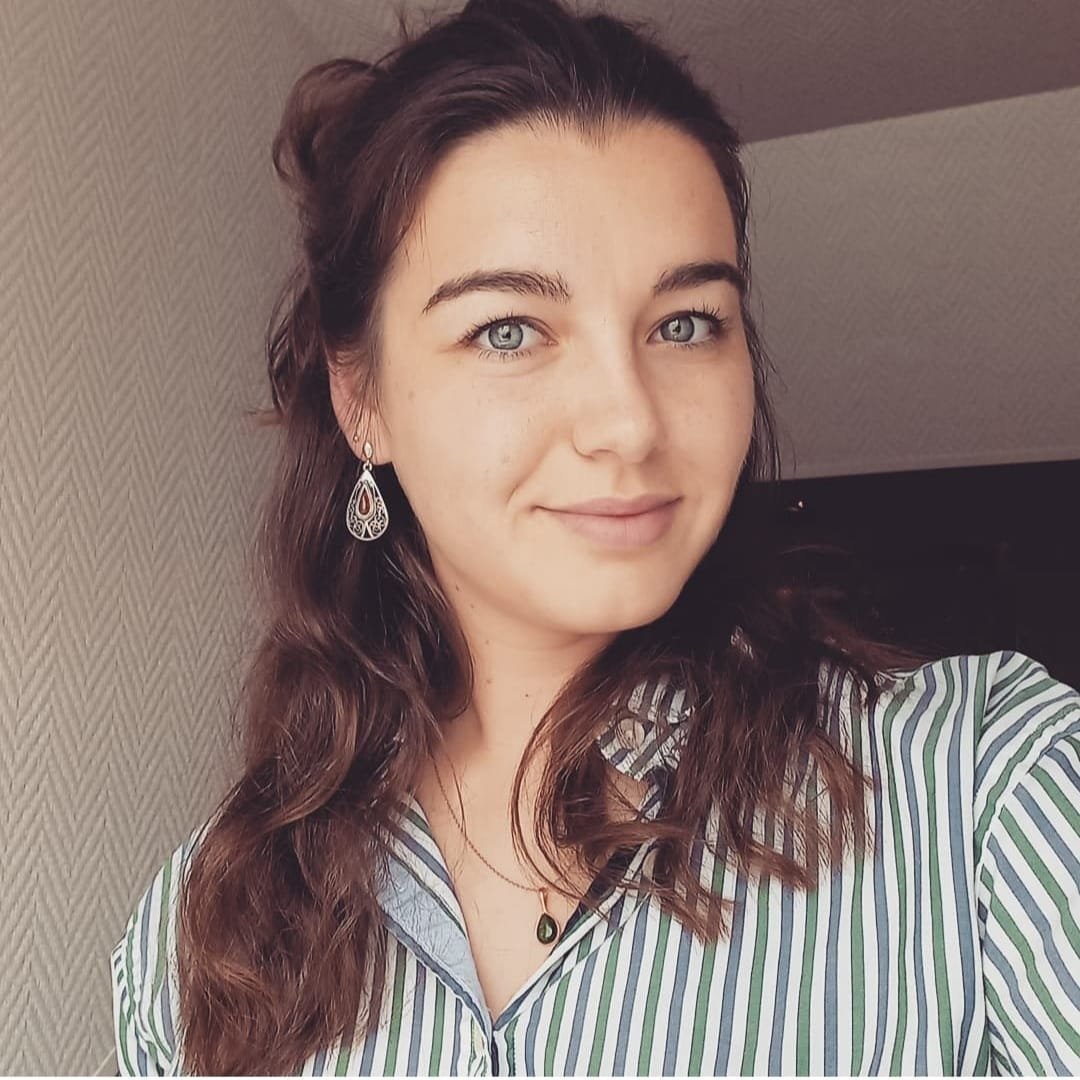 Une jeune femme aux cheveux bouclés, portant des boucles d'oreilles en forme de larme, une chemise à rayures et un collier avec un pendentif noir, sourit légèrement en regardant l'appareil photo.