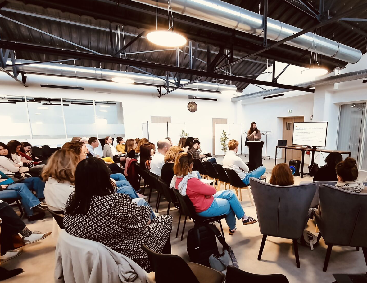 Une salle de conférence moderne où une femme donne une présentation devant un groupe de personnes assises. Il y a un grand écran affichant du texte, des lampes suspendues au plafond, et un décor minimaliste avec quelques plantes.