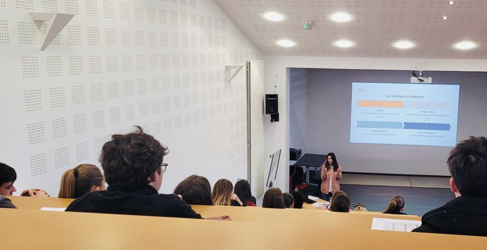 Une salle de classe ou une salle de conférence avec des étudiants ou des participants assis, regardant vers un écran de projection où une femme donne une présentation. L'écran affiche un graphique avec le titre 'Les valeurs en tension'.