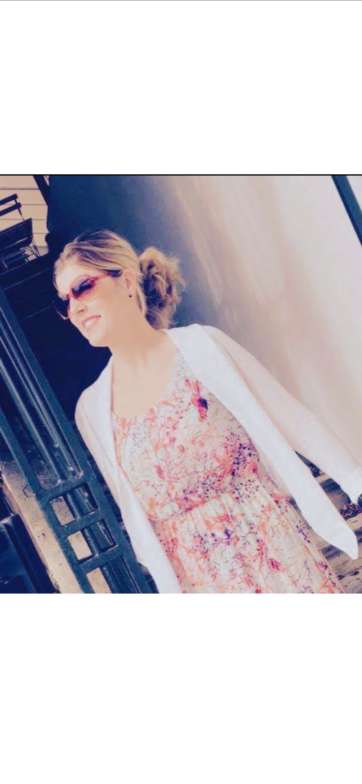 A woman with blonde hair in a messy bun, wearing sunglasses, a floral dress, and a white jacket, standing outside near a railing.
