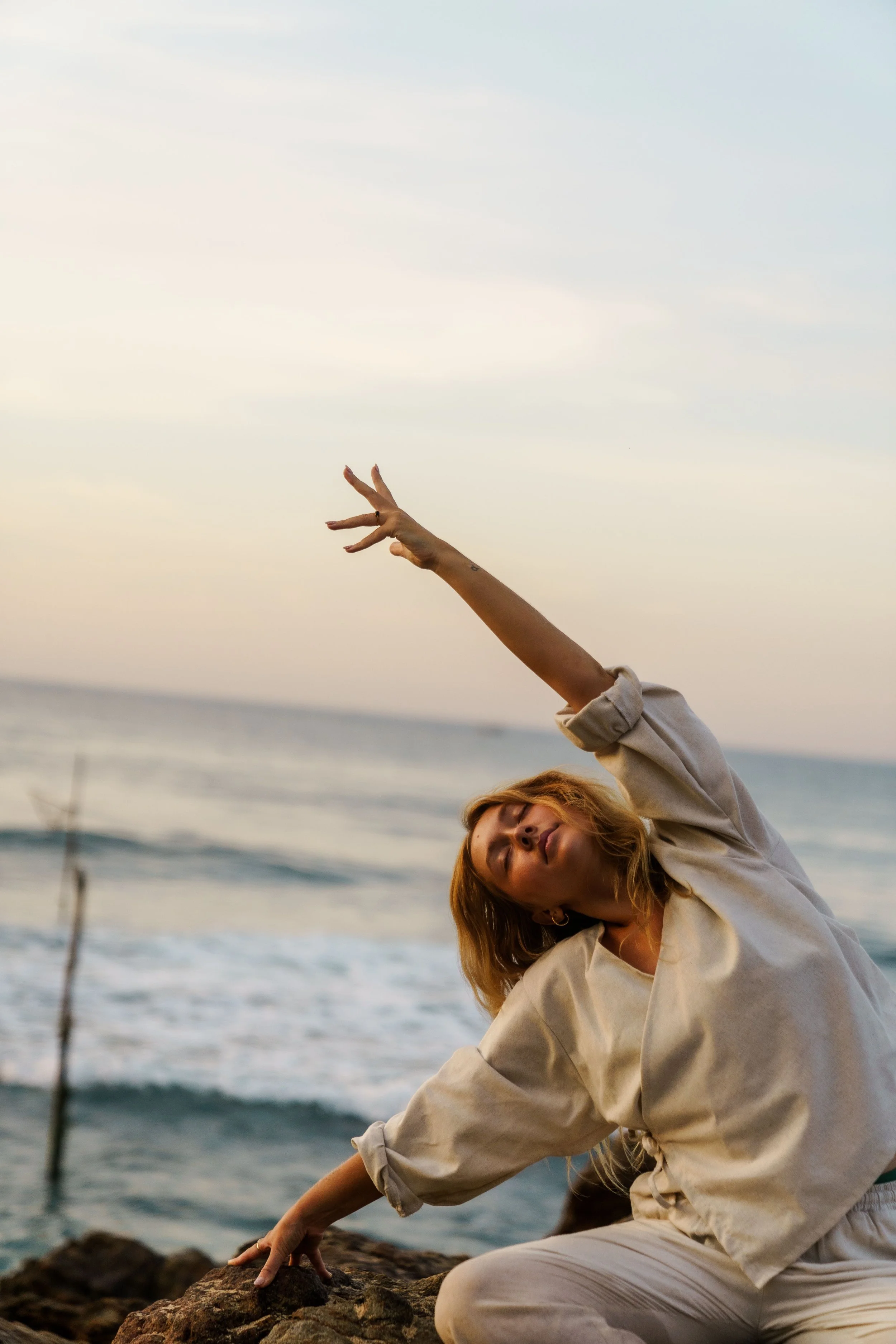 A woman practicing yoga on rocks by the ocean during sunset.