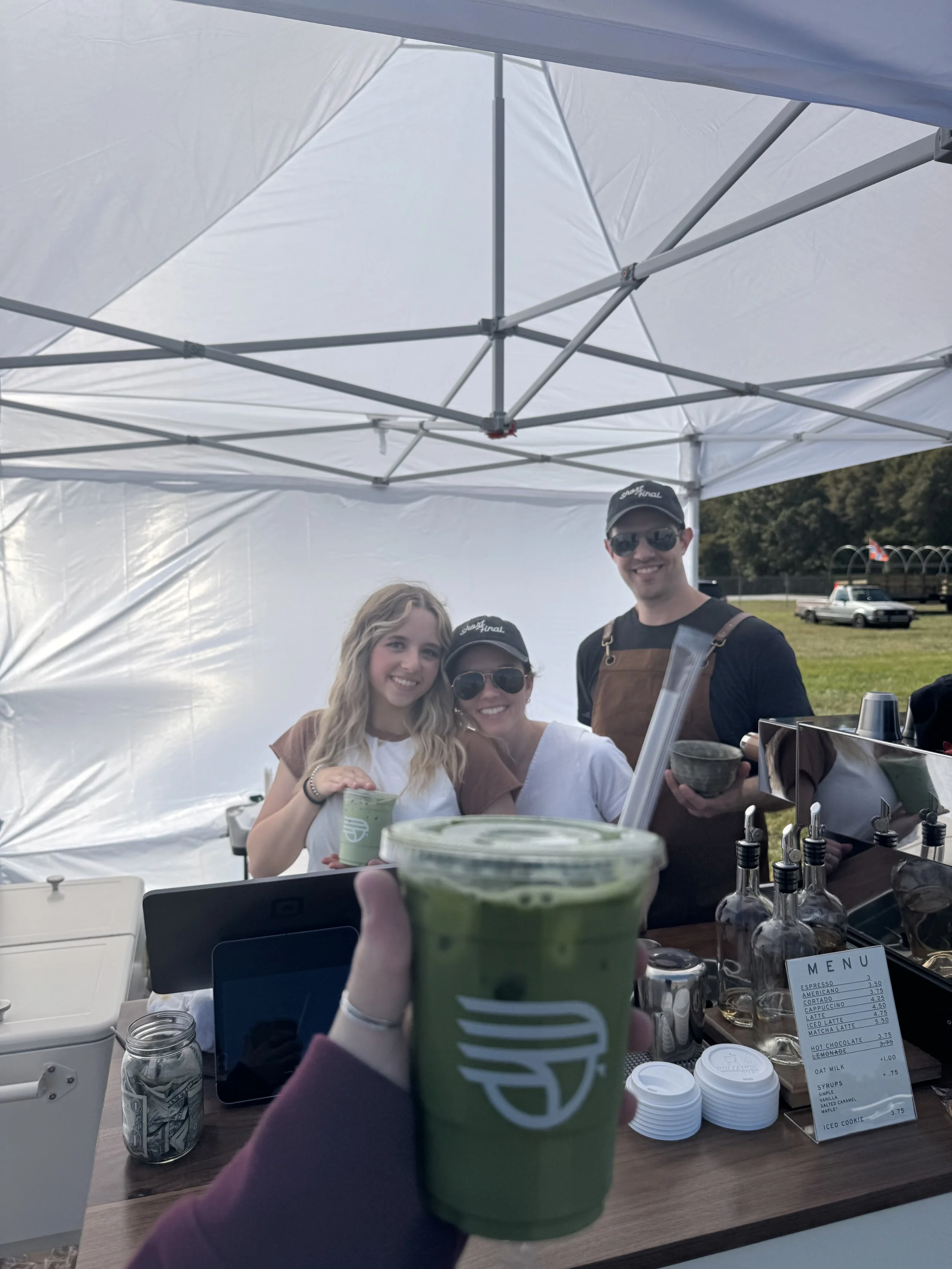 A person holding a green iced matcha latte in front of a coffee stand with three smiling baristas, two women and one man, under a white tent outdoors, with a menu and money on the counter.