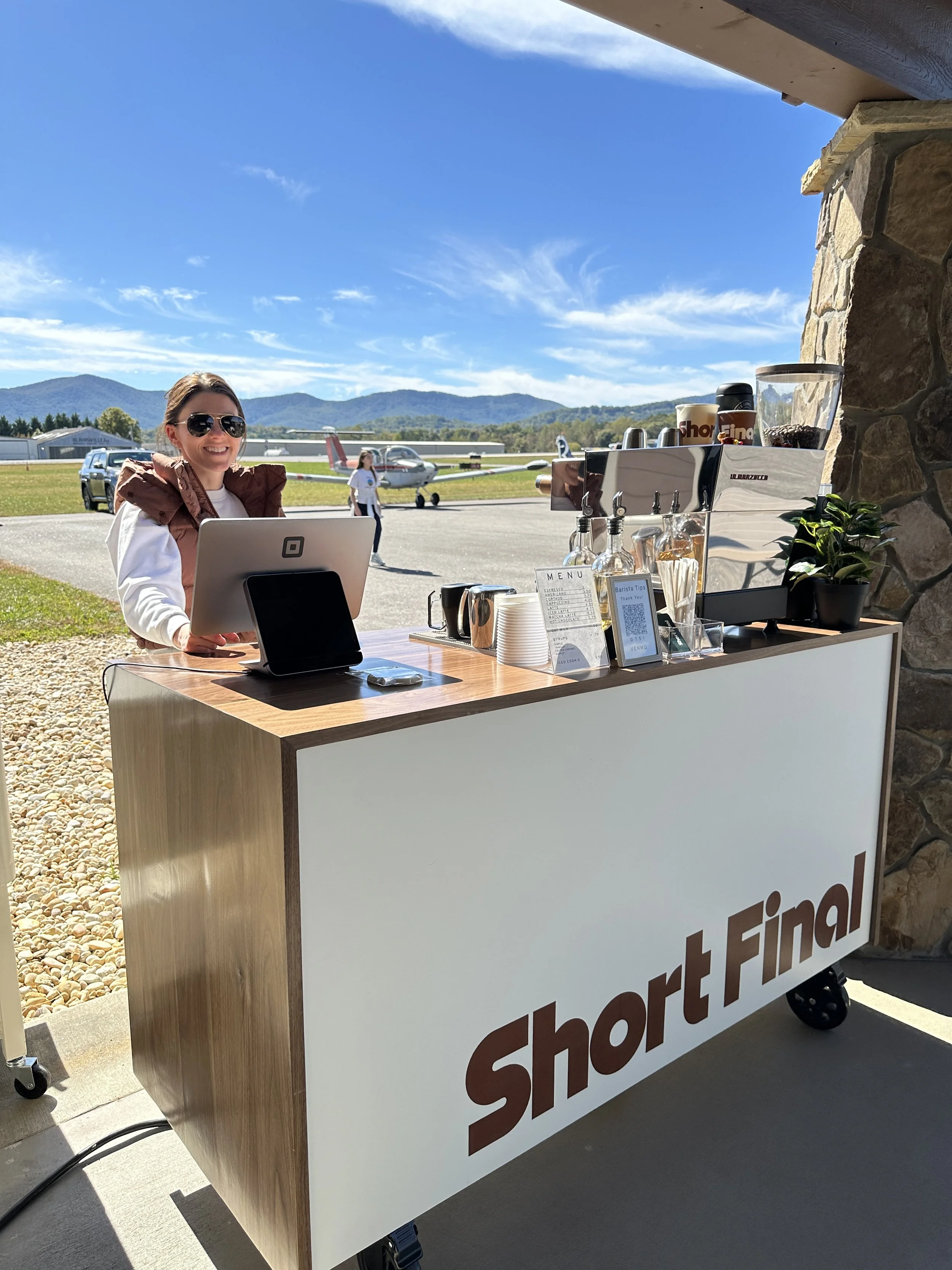 A smiling woman wearing sunglasses stands behind a portable coffee cart labeled "Short Final" at an airport. The cart has a coffee machine, cups, and other supplies on top. Small airplanes and mountains are visible in the background under a blue sky.