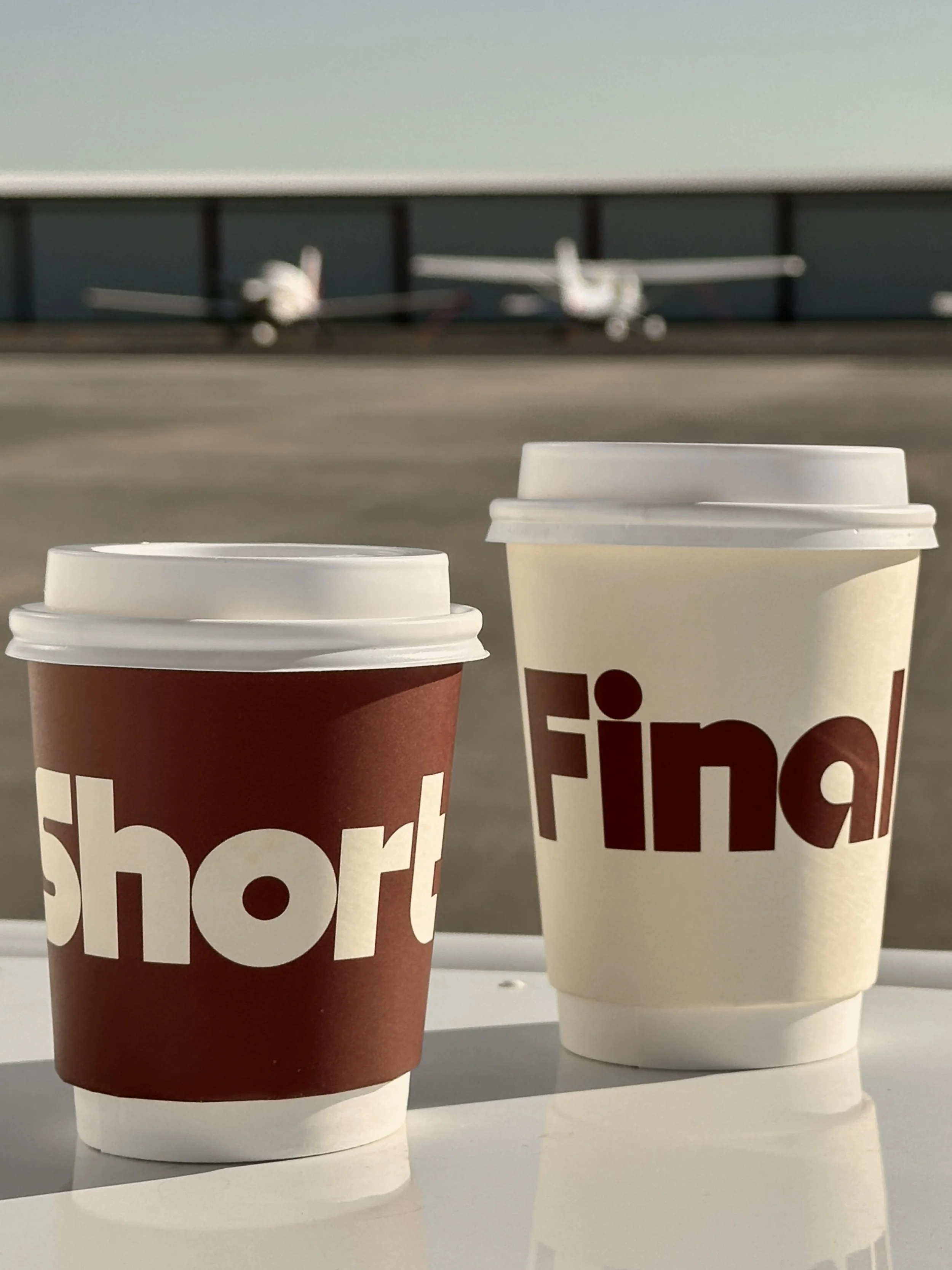 Two paper cups with coffee sit on a white surface, with the words "short" and "final" printed on them. In the background, there are two small airplane models on a rooftop or platform, with a body of water behind them.