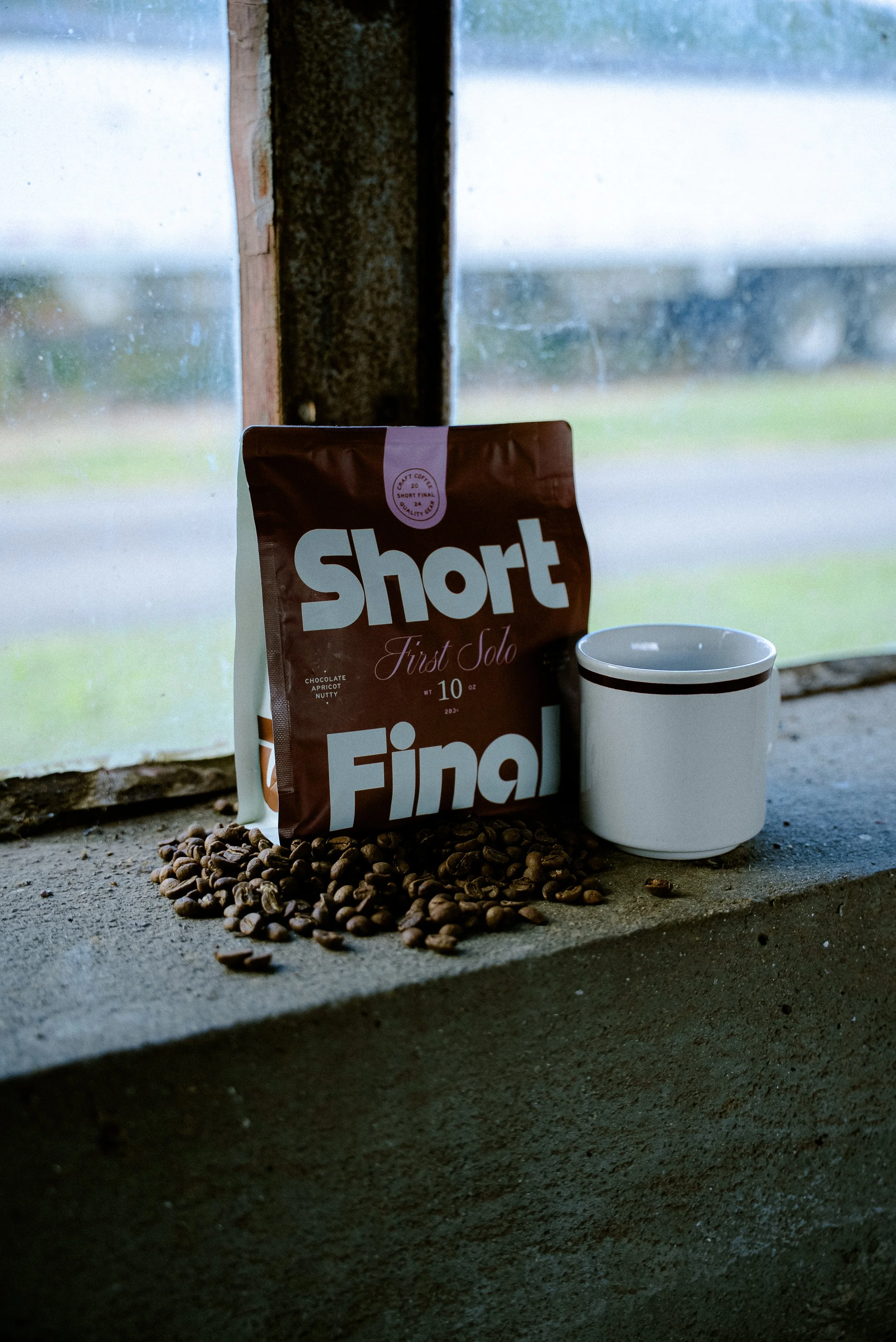 A bag of coffee beans, a pile of coffee beans, and a white mug on a concrete windowsill in front of a rainy window.