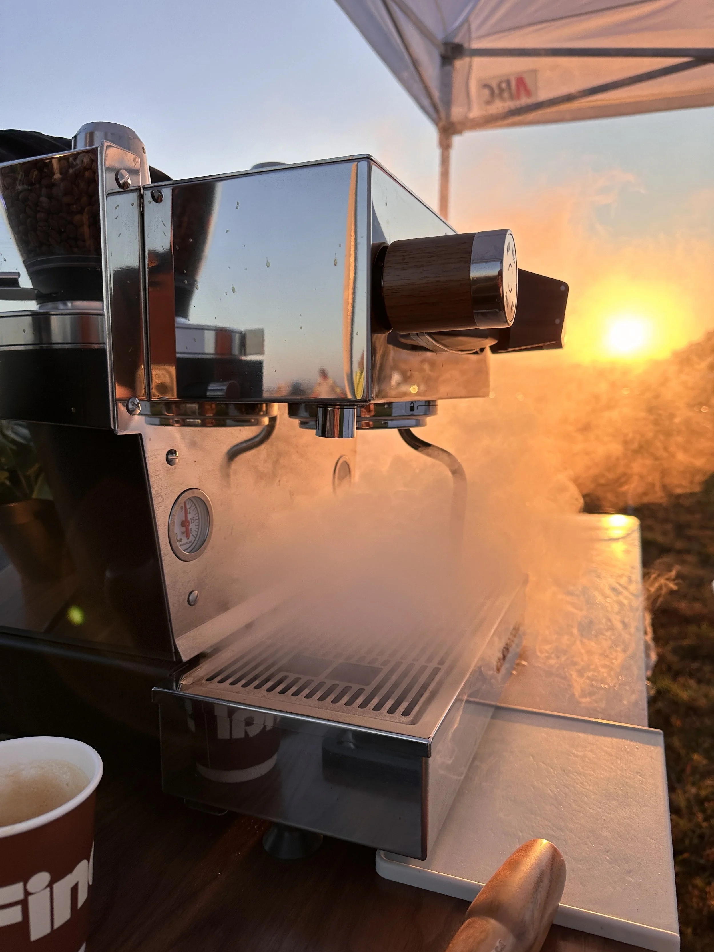 A stainless steel espresso machine with a portafilter attached, steaming milk or water, outdoors during sunset with a tent overhead and a coffee cup nearby.