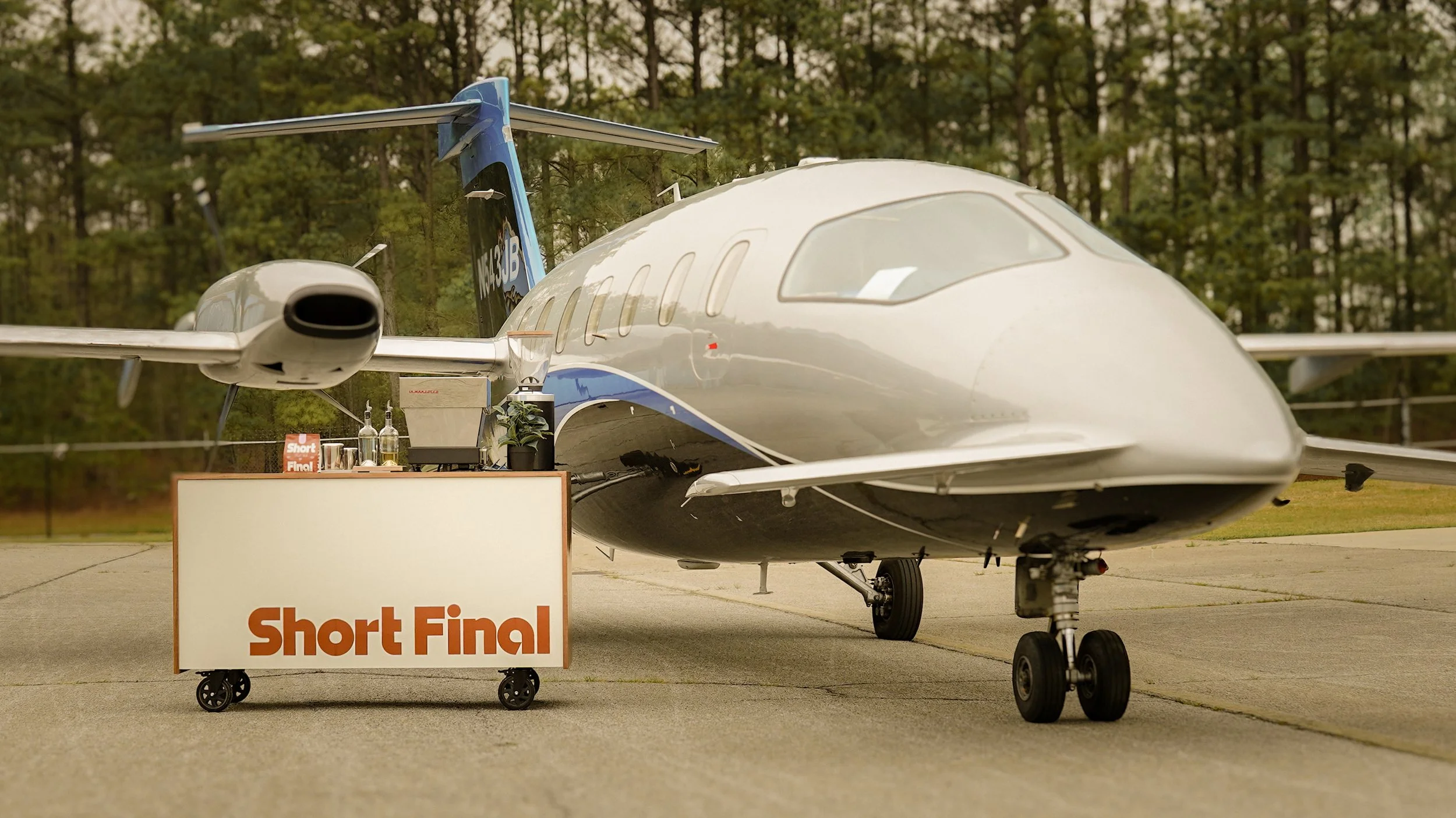A small private jet parked outdoors next to a cart with a sign that reads 'Short Final'. The cart has bottles and a coffee setup on top. The area is surrounded by trees.