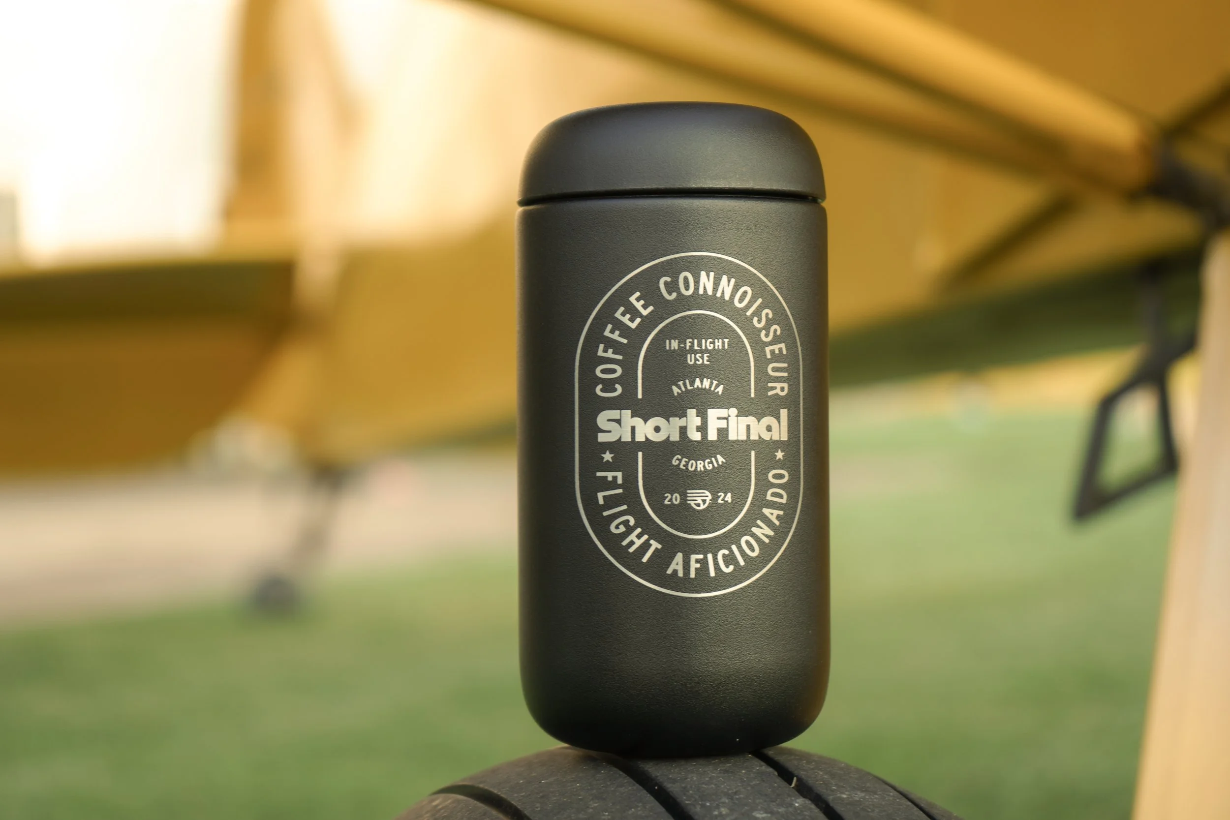 Black insulated tumbler with white circular graphics and text related to coffee, aviation, and a flight club, placed outdoors on a tire, with a blurred yellow playground structure in the background.