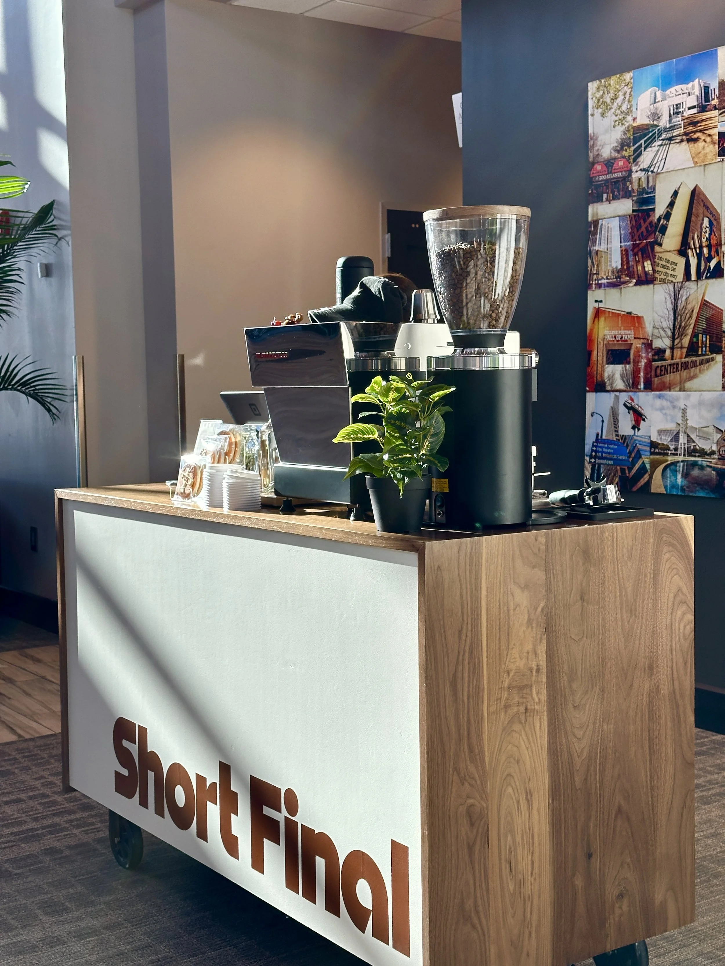 Coffee counter in a cafe with a white front panel featuring the text 'short Final' in brown, a coffee grinder, a coffee machine, cups, and a potted plant on top, with photos of buildings on the wall behind.