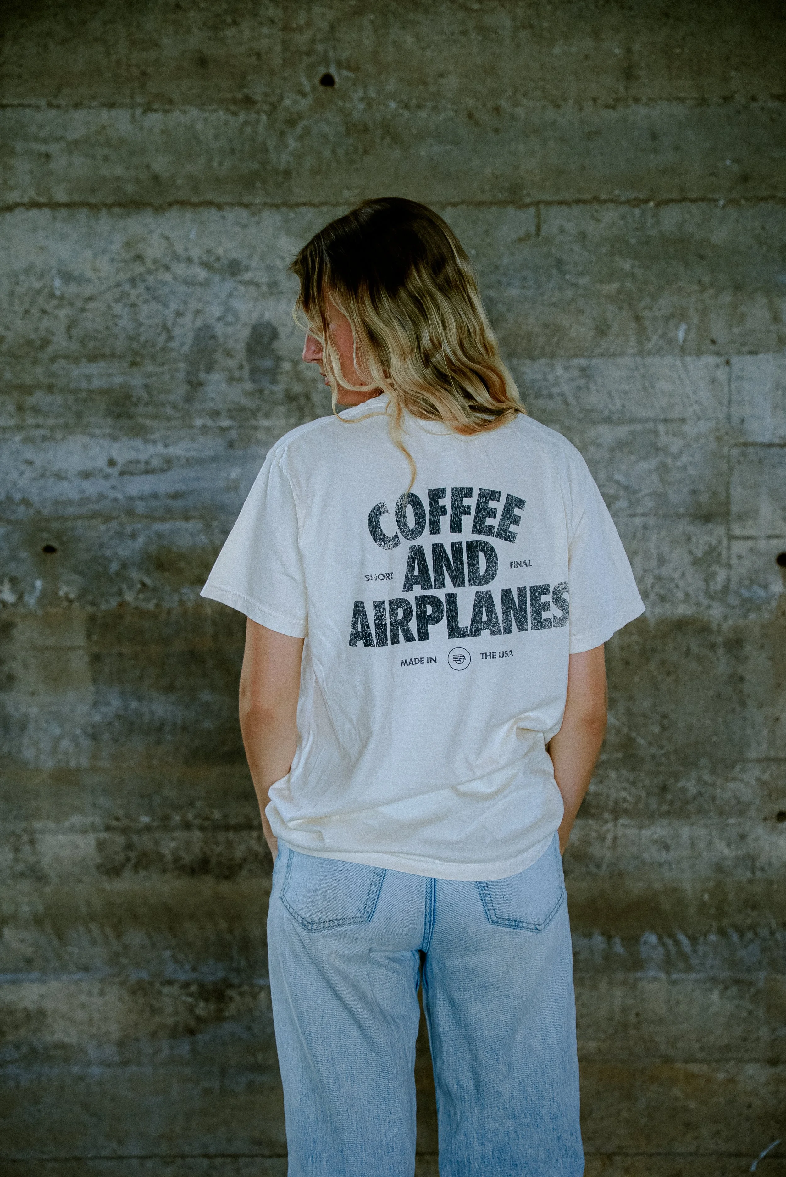 A woman with wavy blonde hair standing with her back to the camera, wearing a white T-shirt with black text and light blue jeans, in front of a concrete wall.