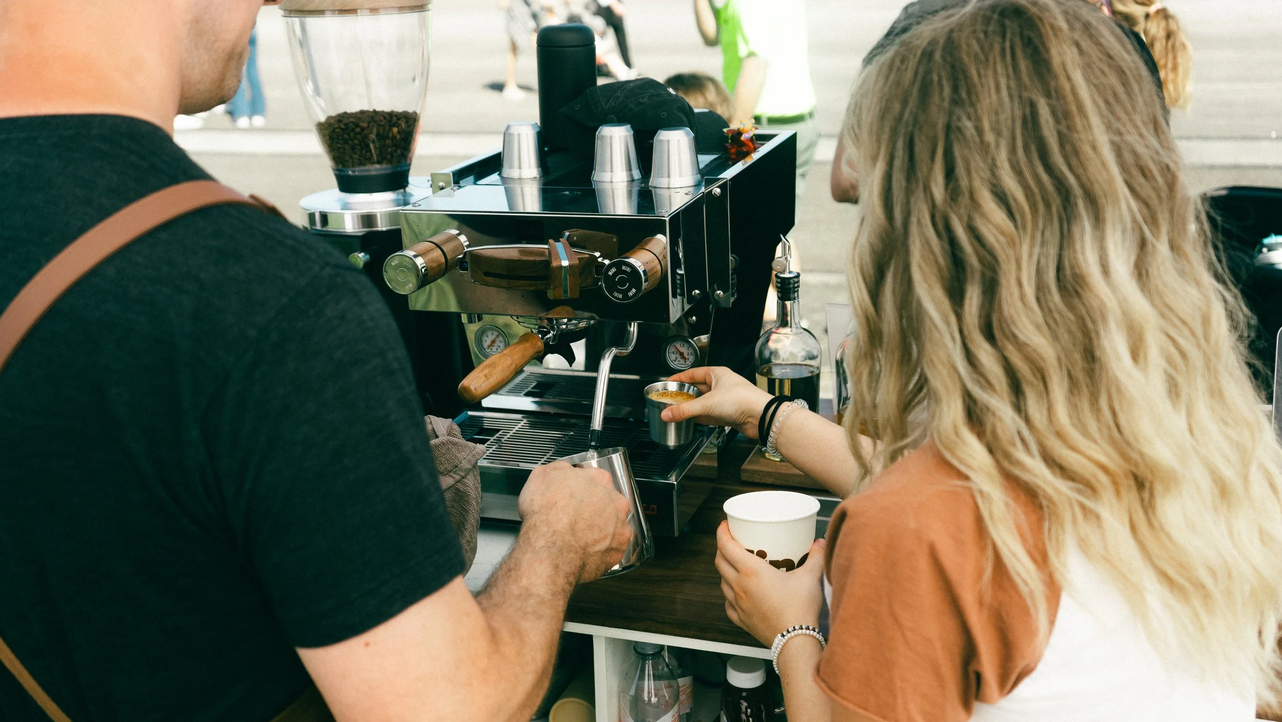 A barista making espresso at an outdoor coffee stand with a woman getting a coffee drink.
