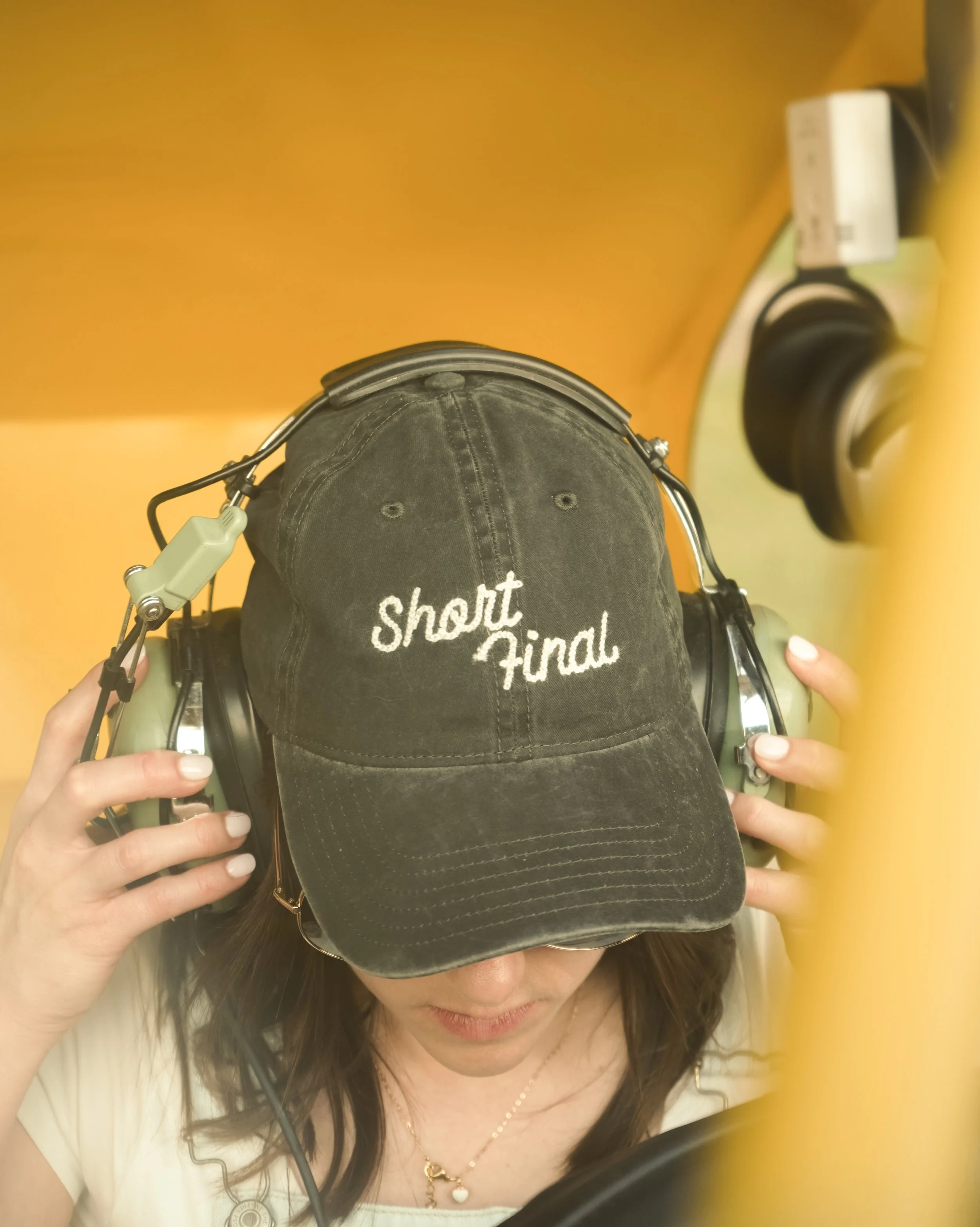 A person wearing a black baseball cap with the words 'Short Final' embroidered in white, and large green headphones, appears to be in an airplane cockpit or control room.