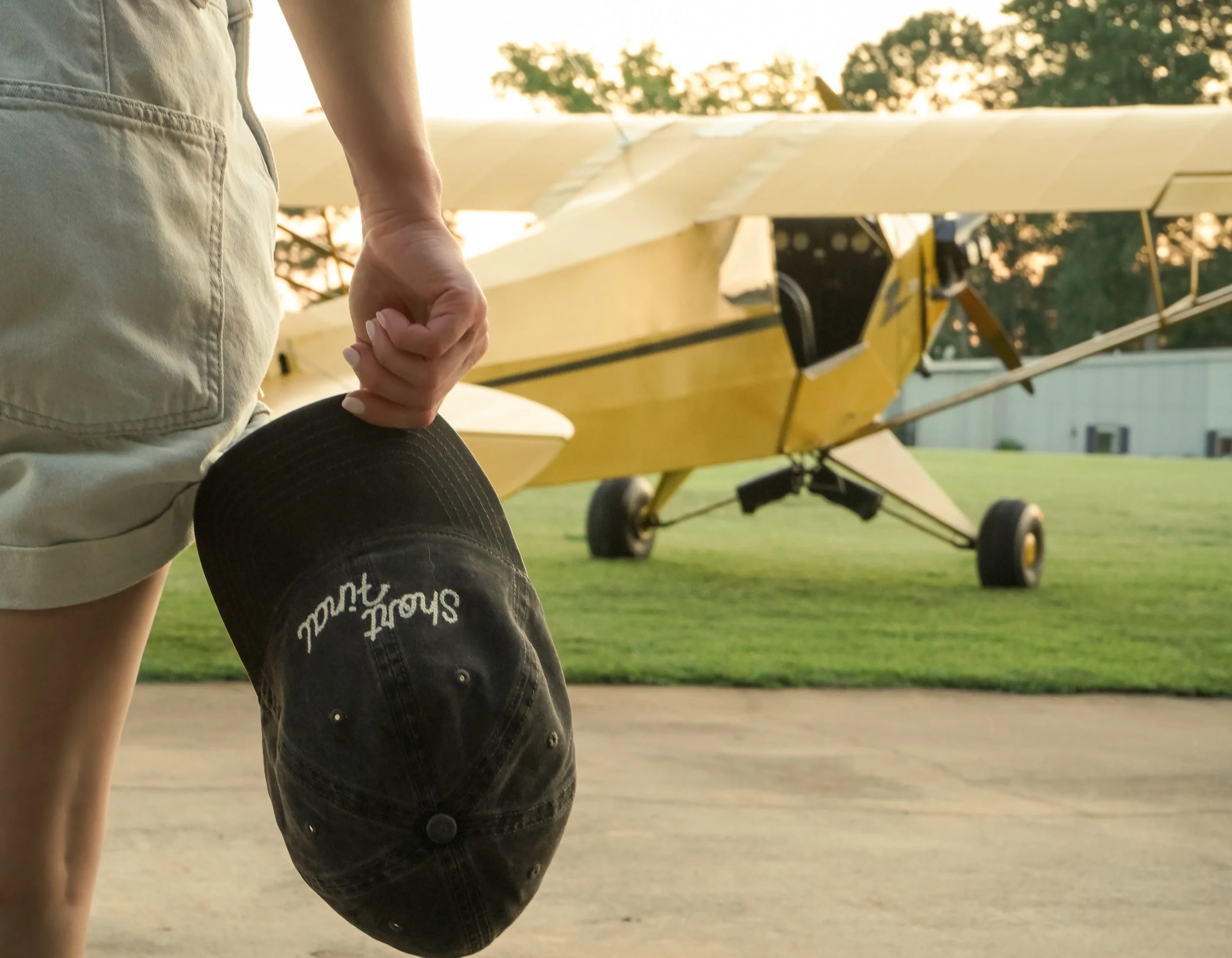 A person is holding a black baseball cap with embroidered text in front of a small yellow airplane parked on a grassy field during sunset.