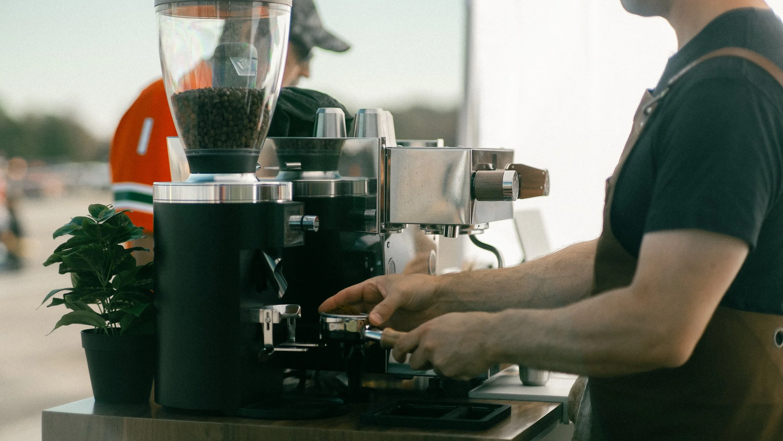 Barista preparing coffee behind a professional espresso machine outdoors, with a small plant next to the machine. Airplane Fly-In event. Pilots and aviation enthusiasts enjoying Short Final Coffee craft beverages