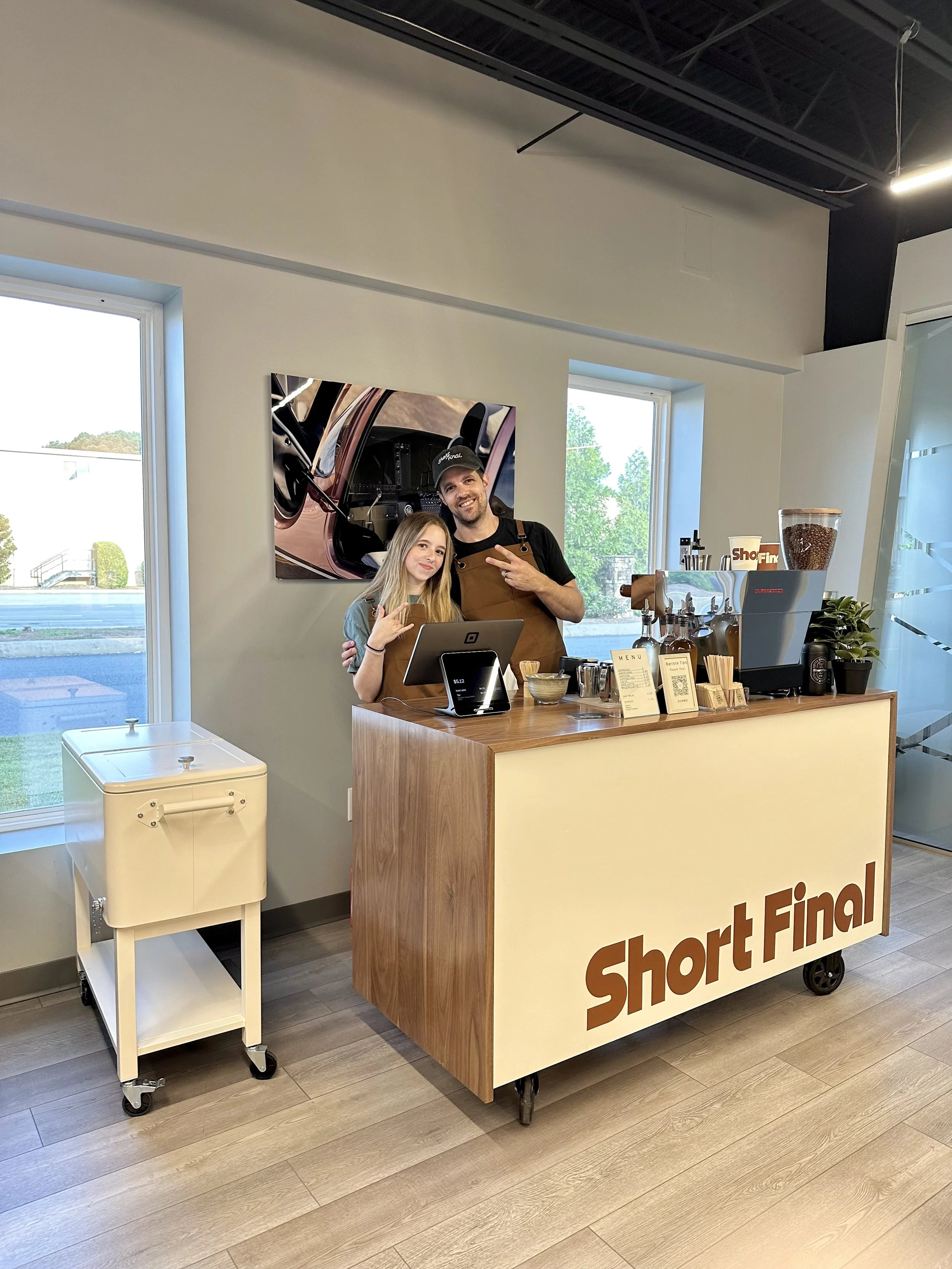 Two people in a coffee shop behind a counter with the sign 'Short Final'. One is a young girl, and the other is a man wearing a brown apron and cap, both making peace signs, smiling. The coffee shop has two large windows, a colorful poster on the wal