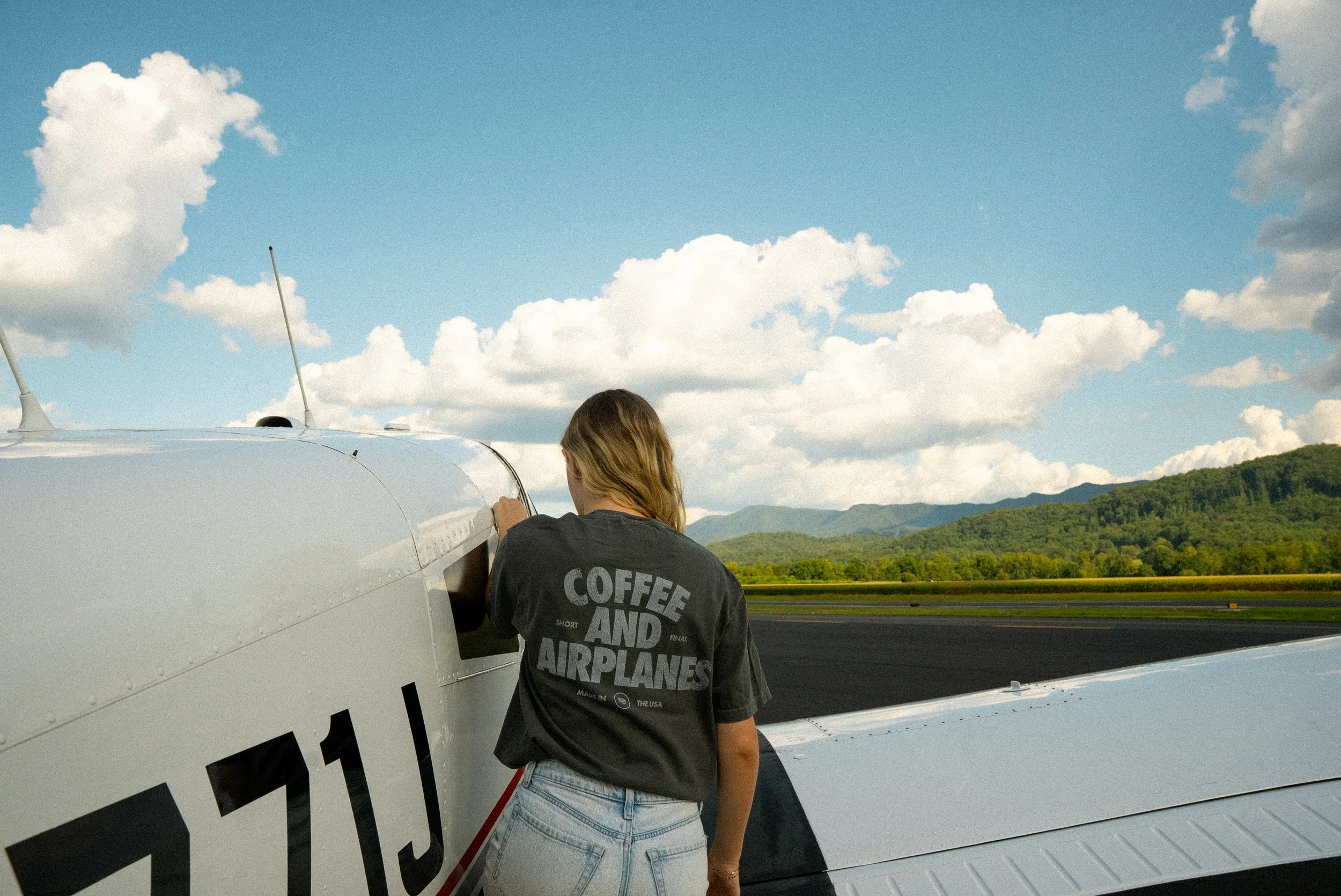 A woman in a black t-shirt with the words "Coffee and Airplanes" stands next to a small white airplane on a runway, with mountains and clouds in the background.