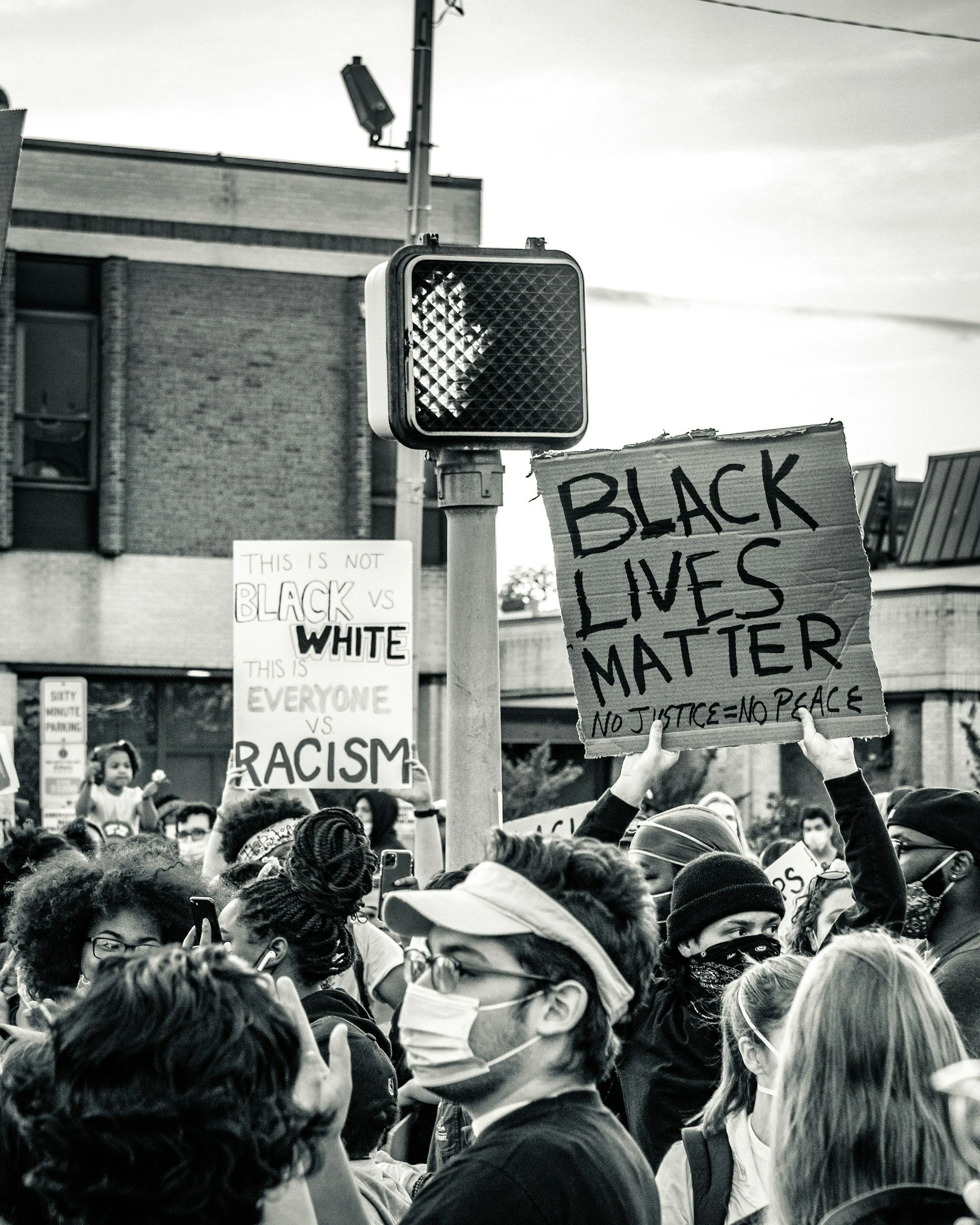 Crowd of protesters holding signs at a Black Lives Matter rally, wearing masks, with one sign reading 'Black Lives Matter' and another quoting 'No Justice = No Peace'.