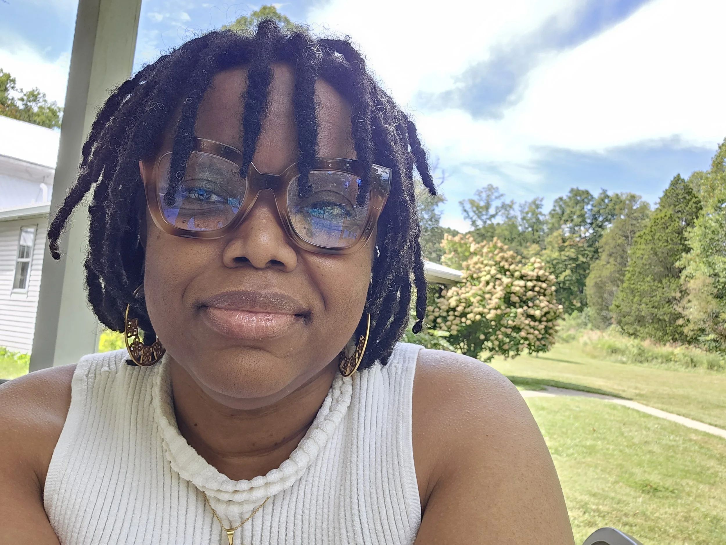 A woman with dreadlocks wearing sunglasses, earrings, a white sleeveless top, and a necklace, outdoors with trees and a garden in the background on a sunny day.