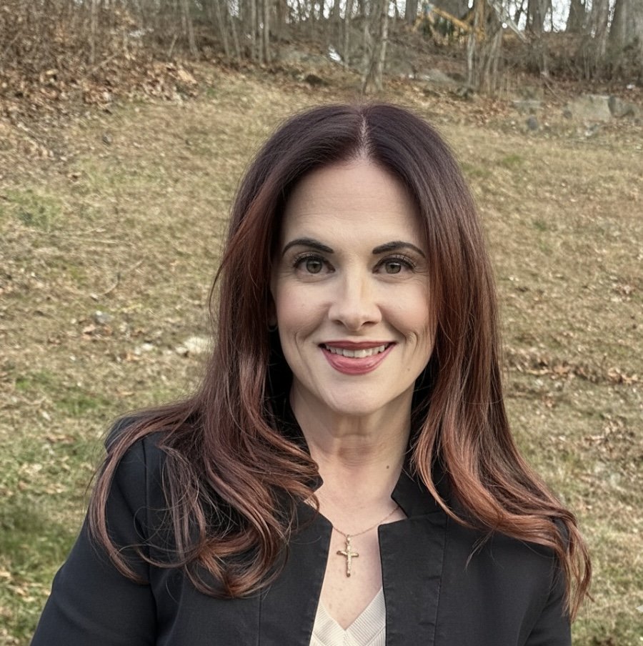 A woman with long, wavy brown hair and light skin, smiling outdoors, wearing a black jacket and a cross necklace, with a grassy hill and trees in the background.