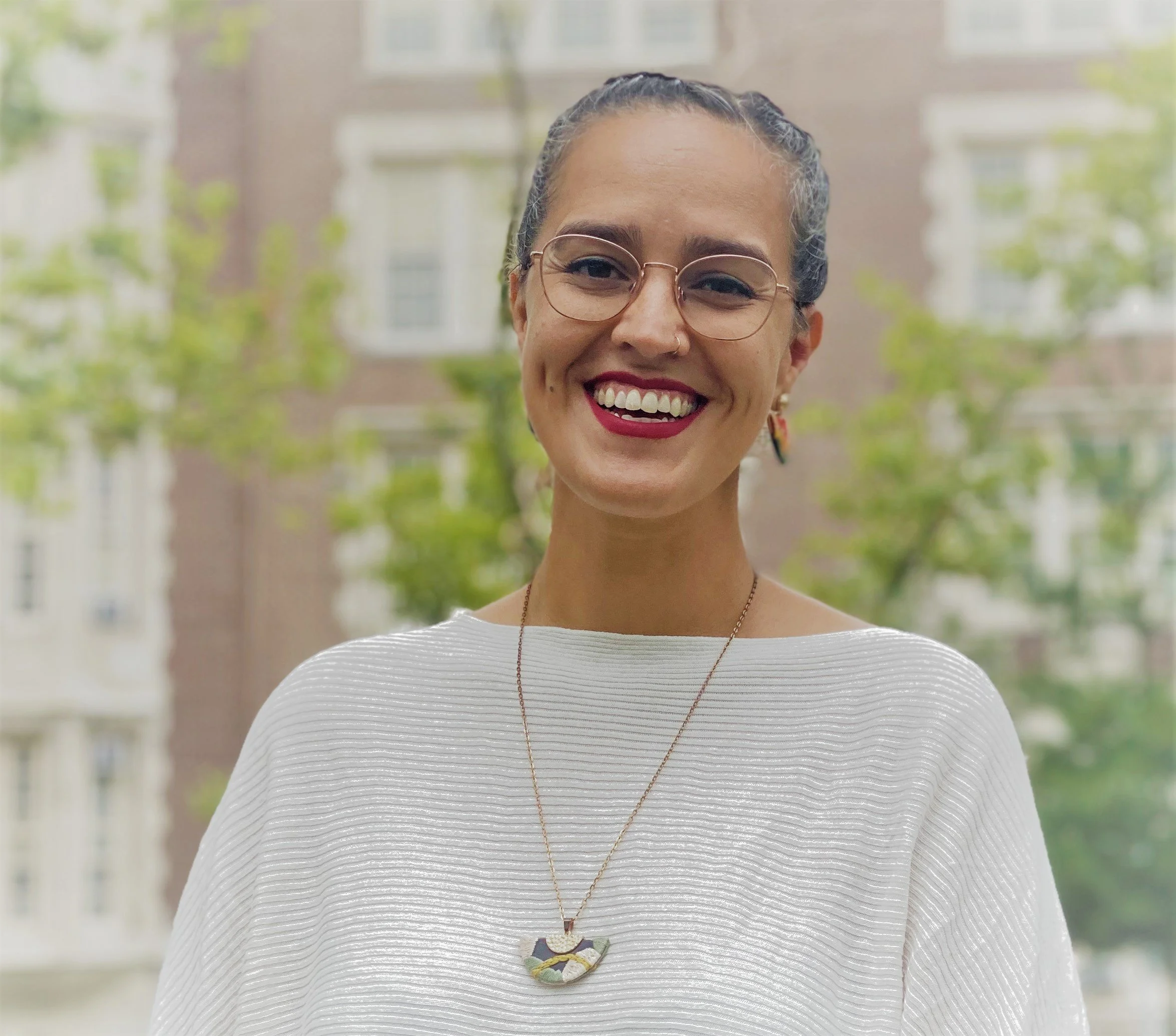 Smiling woman with glasses and earrings, wearing a white top with textured stripes and a colorful necklace, standing outdoors with trees and buildings in the background.