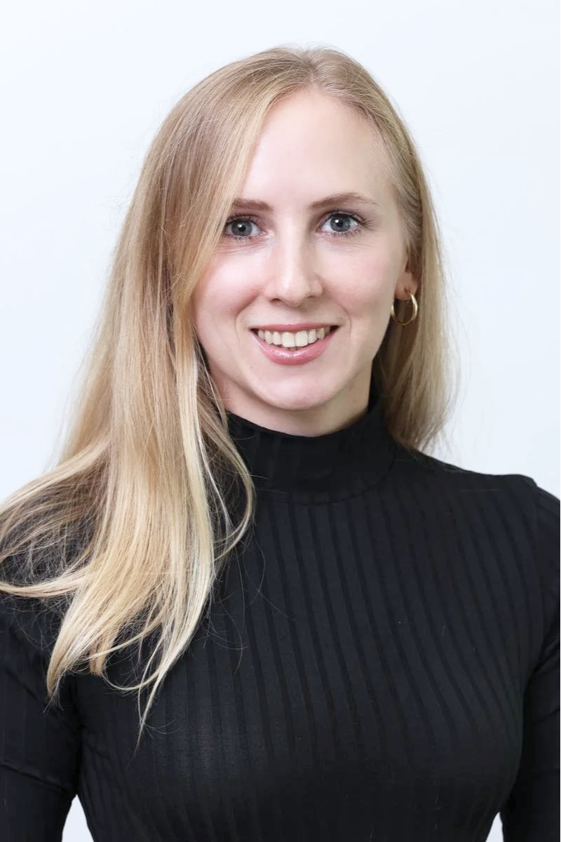 Porträt von Anja Felicitas Beyerbach mit langem blondem Haar, schwarzen Rollkragenpullover und goldenen Ohrringen, vor hellem Hintergrund.