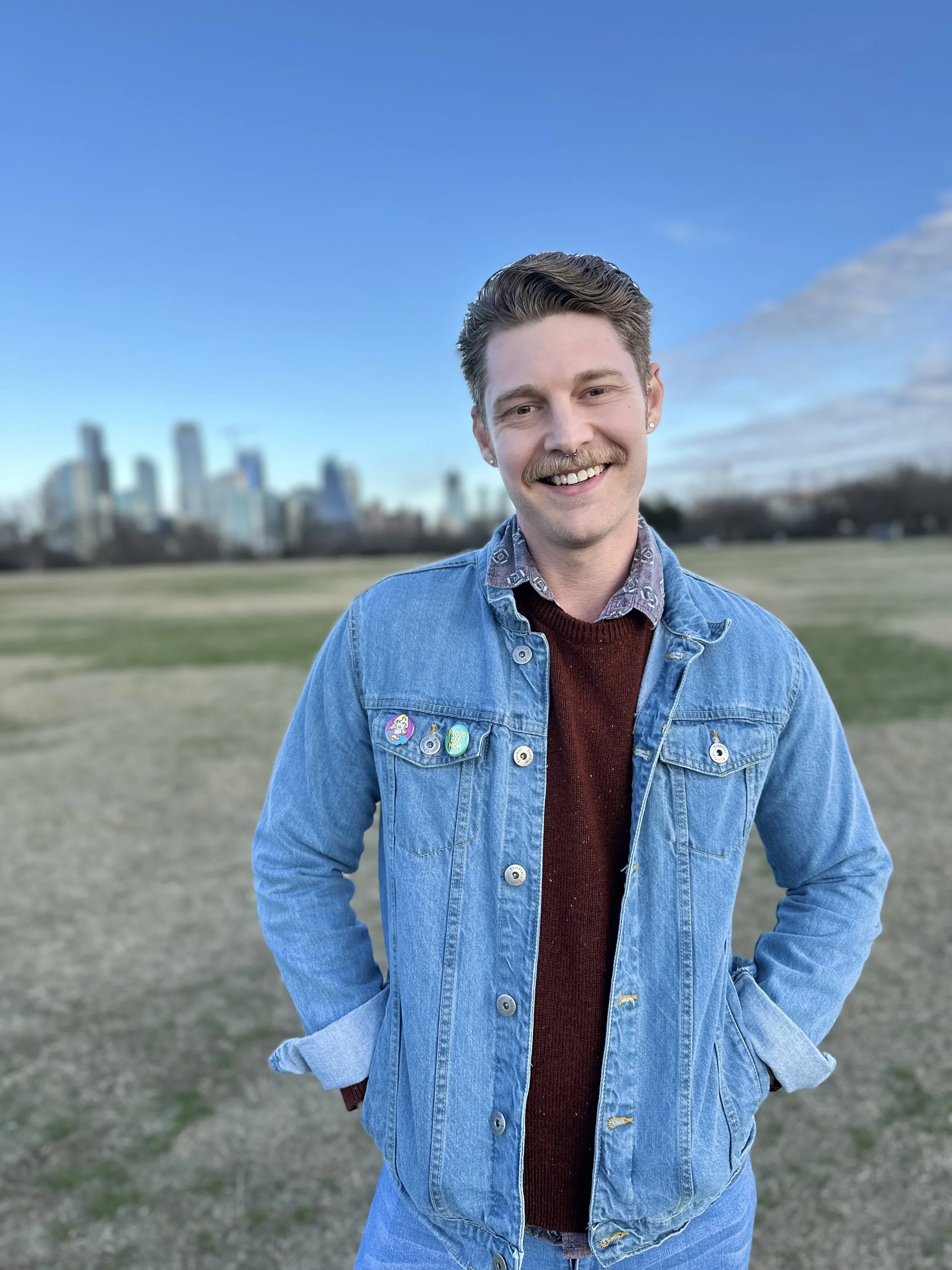 A young man standing outdoors in a park with a city skyline in the background. He has short brown hair, a mustache, and is smiling. He is wearing a denim jacket with pins and a maroon sweater underneath.