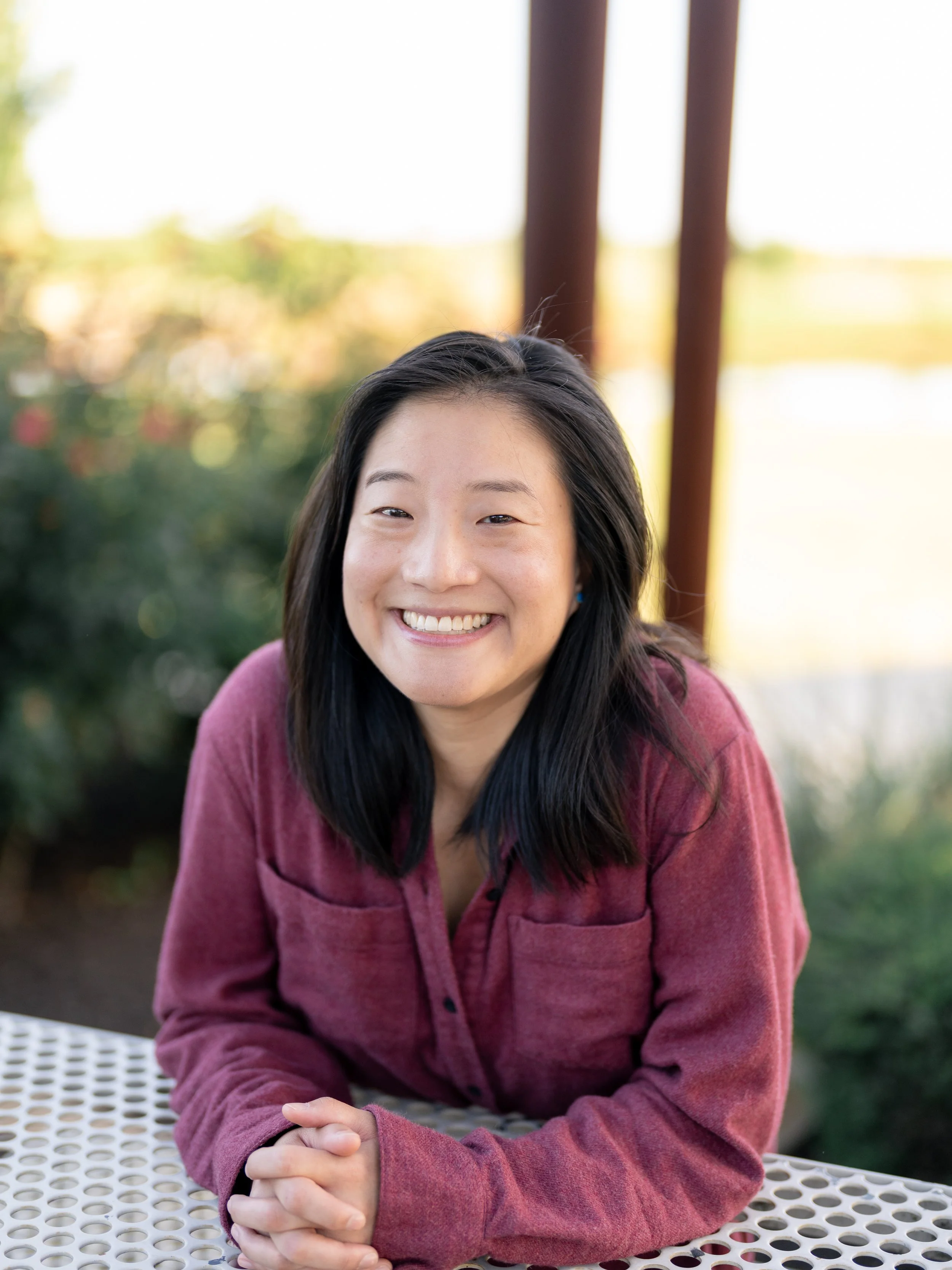 Smiling woman with black hair sitting at a perforated table outdoors, wearing a red long sleeve shirt, with a blurred green and brown background and wooden beams.